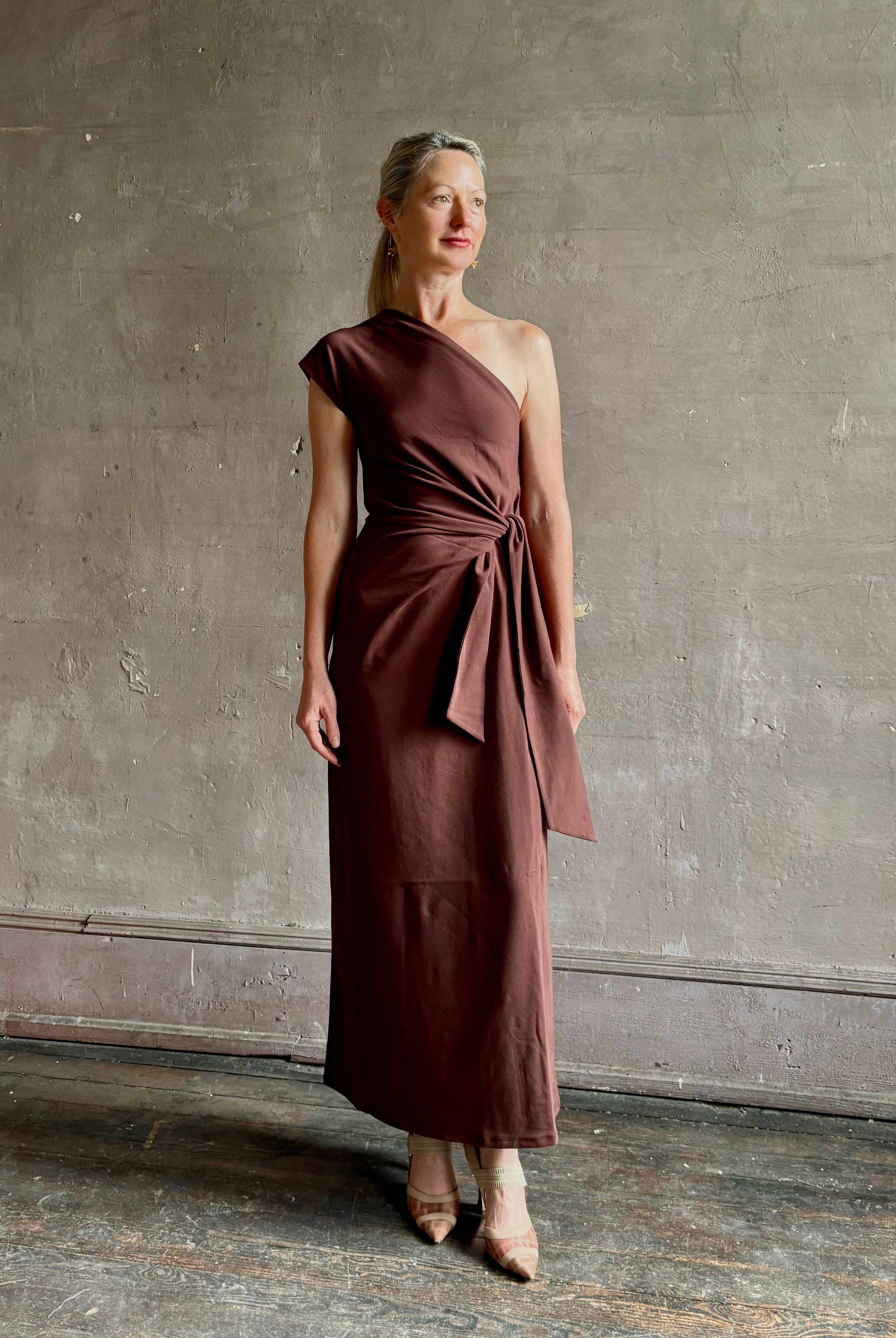 Mature woman wearing Apiece Apart brown comfy jersey cotton maxi dress one shoulder twist tie waist stretch