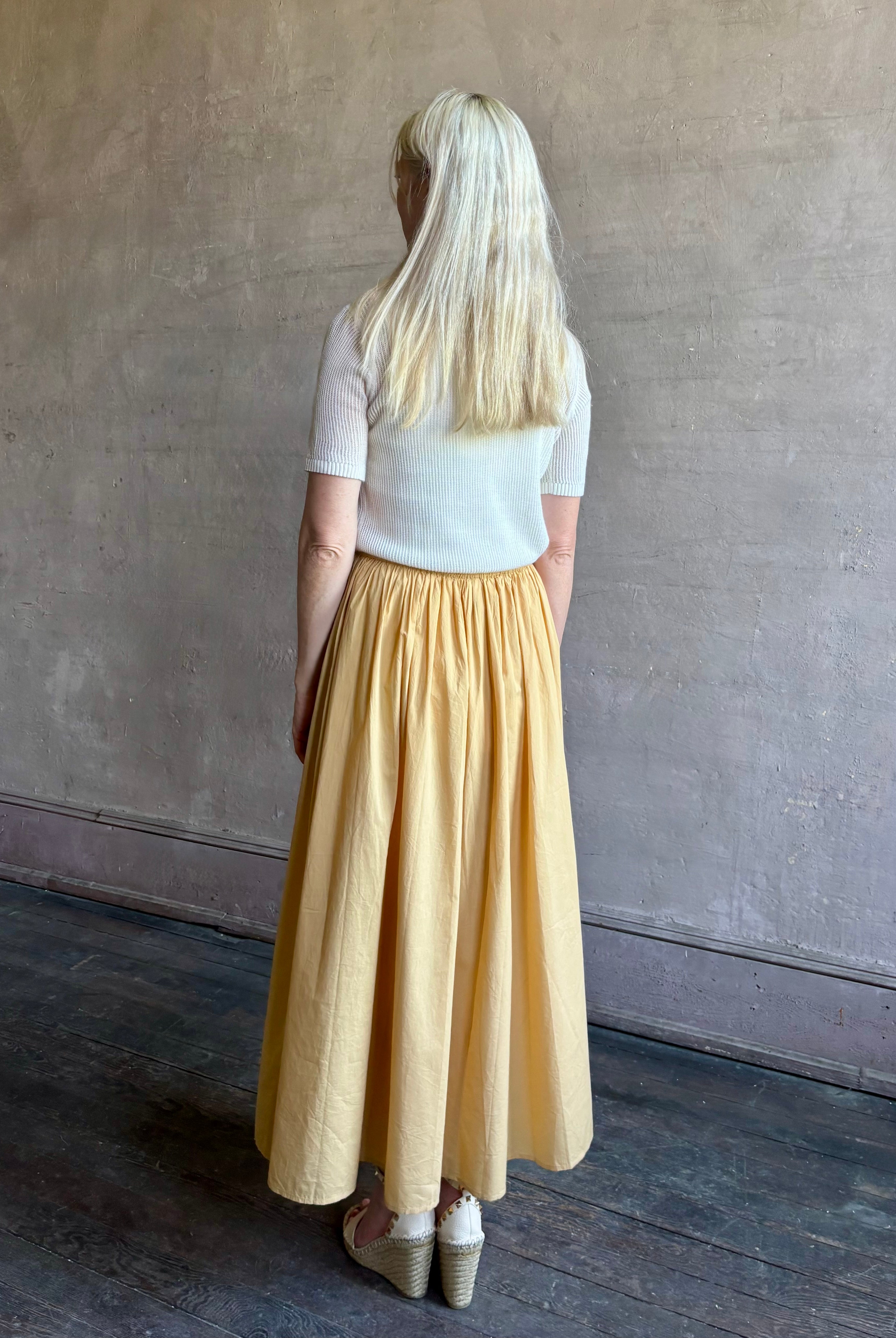 Mature woman wearing the Hanne Tie Maxi Skirt from Apiece Apart rendered in a butter yellow hue and crafted from 100% organic cotton with adjustable drawstring and elastic waistband