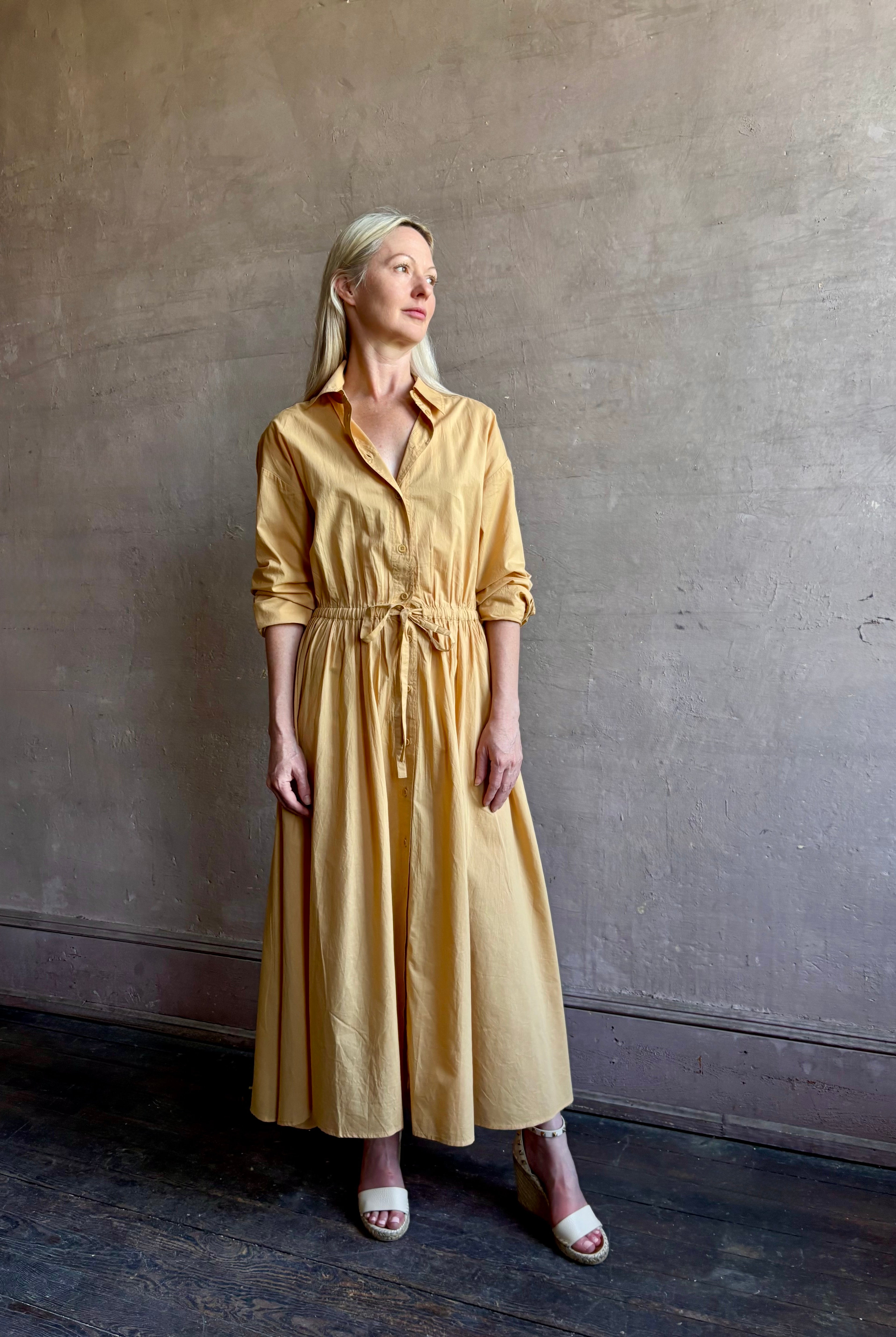 Image of mature woman wearing the Priska Shirt Dress from Apiece Apart rendered in a butter yellow and crafted from 100% organic cotton featuring long sleeves, collared button front and adjustable drawstring waistband
