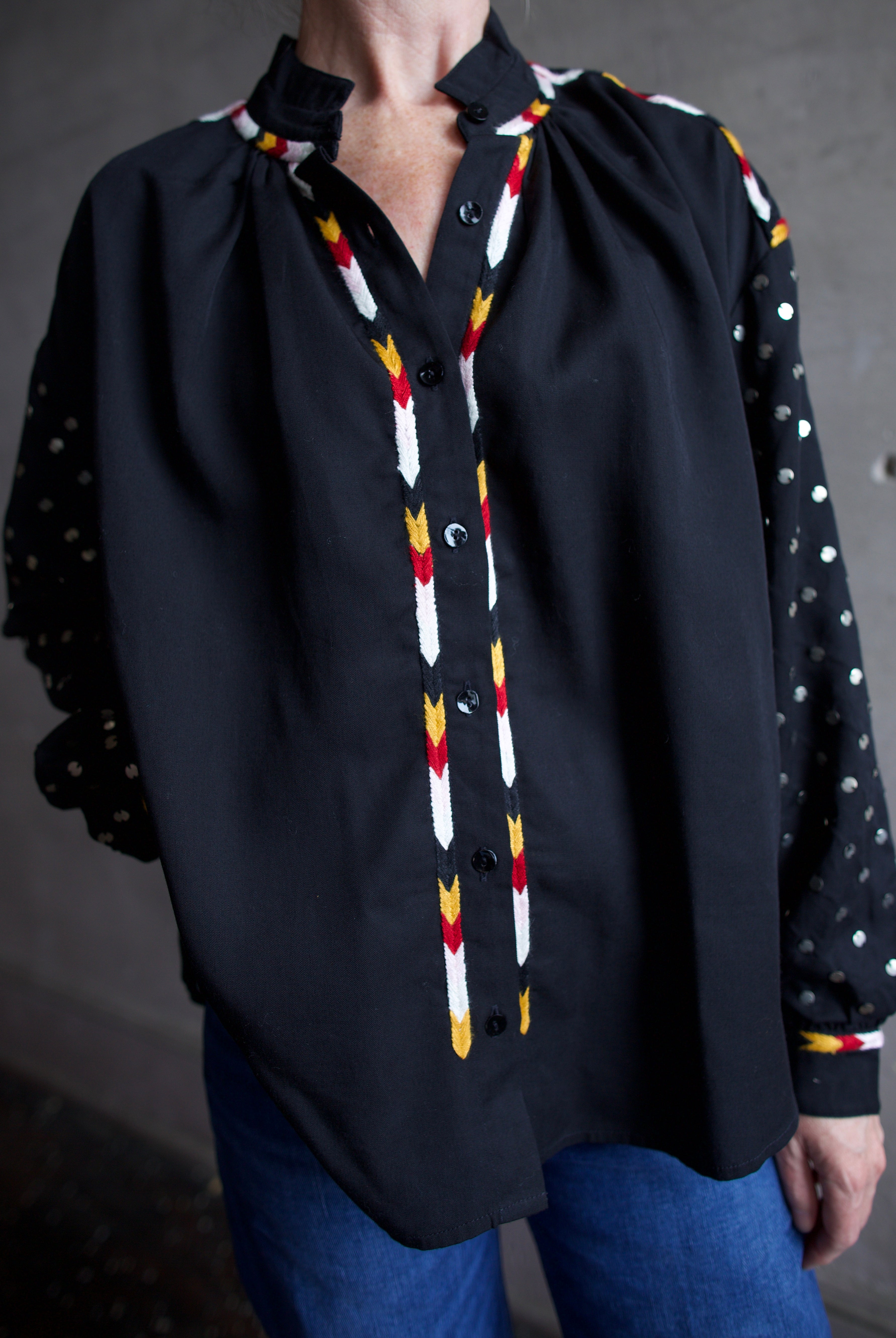 Image featuring a woman wearing the Alix of Bohemia Kiki Stardust button front black shirt with red, white, yellow, & black embroidery and metal discs on the long sleeves.