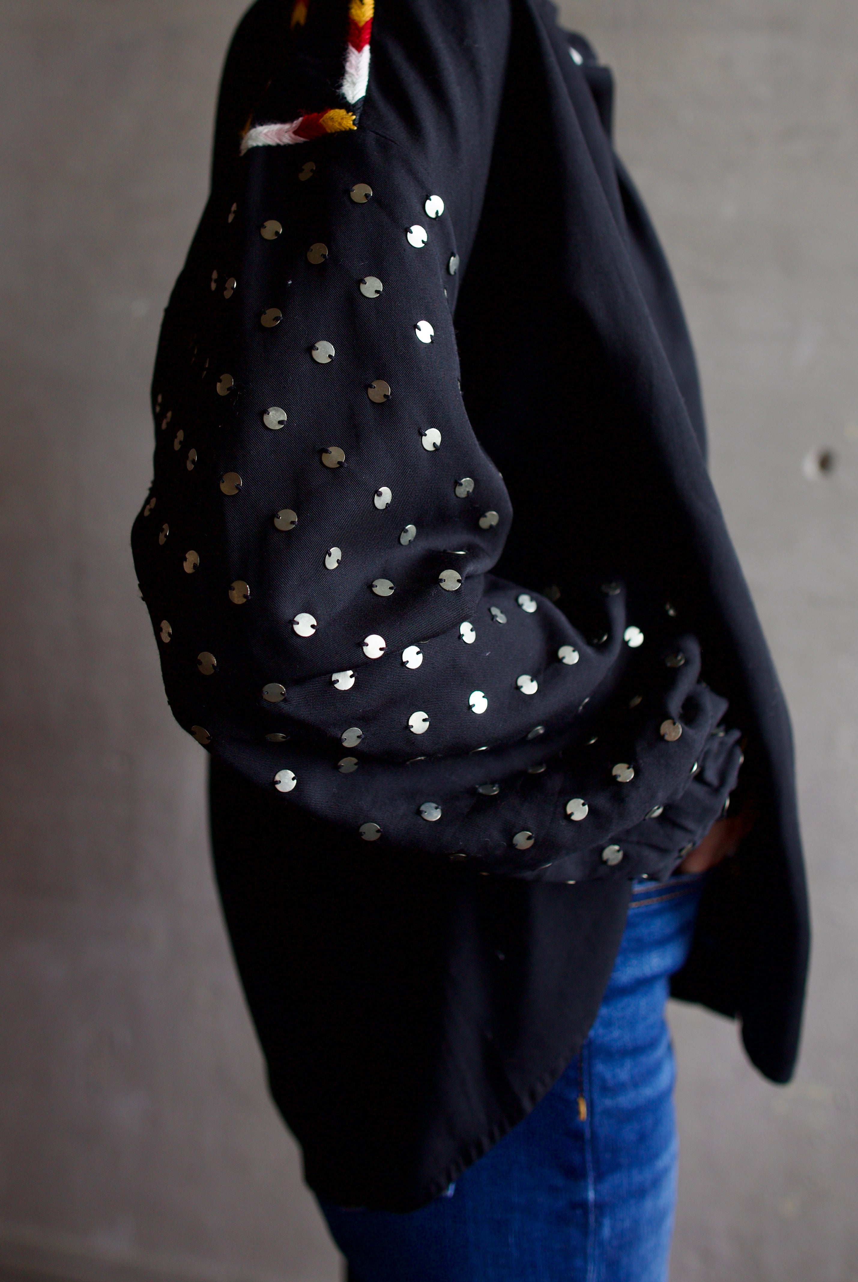 Image featuring a woman wearing the Alix of Bohemia Kiki Stardust button front black shirt with red, white, yellow, & black embroidery and metal discs on the long sleeves.