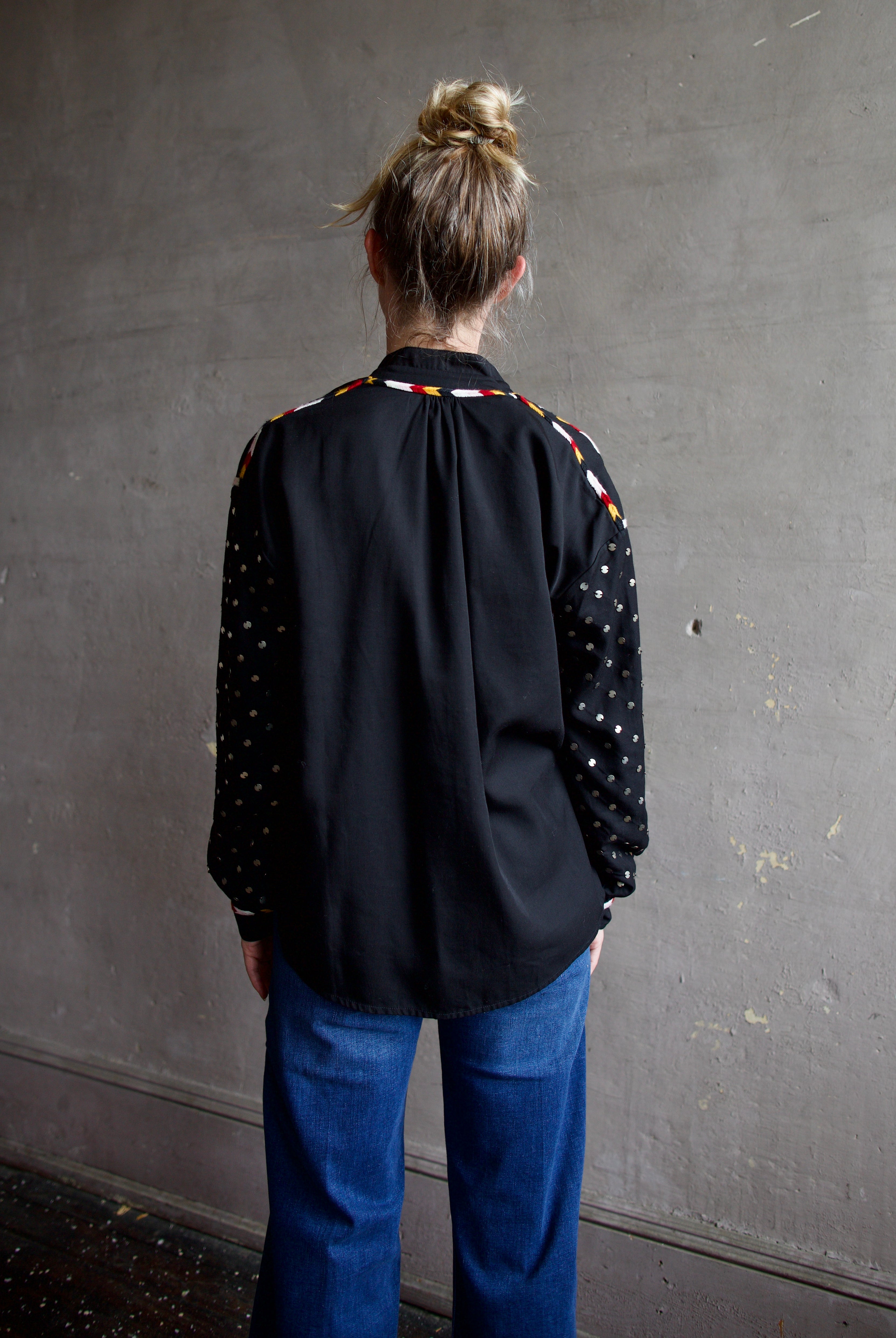 Image featuring a woman wearing the Alix of Bohemia Kiki Stardust button front black shirt with red, white, yellow, & black embroidery and metal discs on the long sleeves.