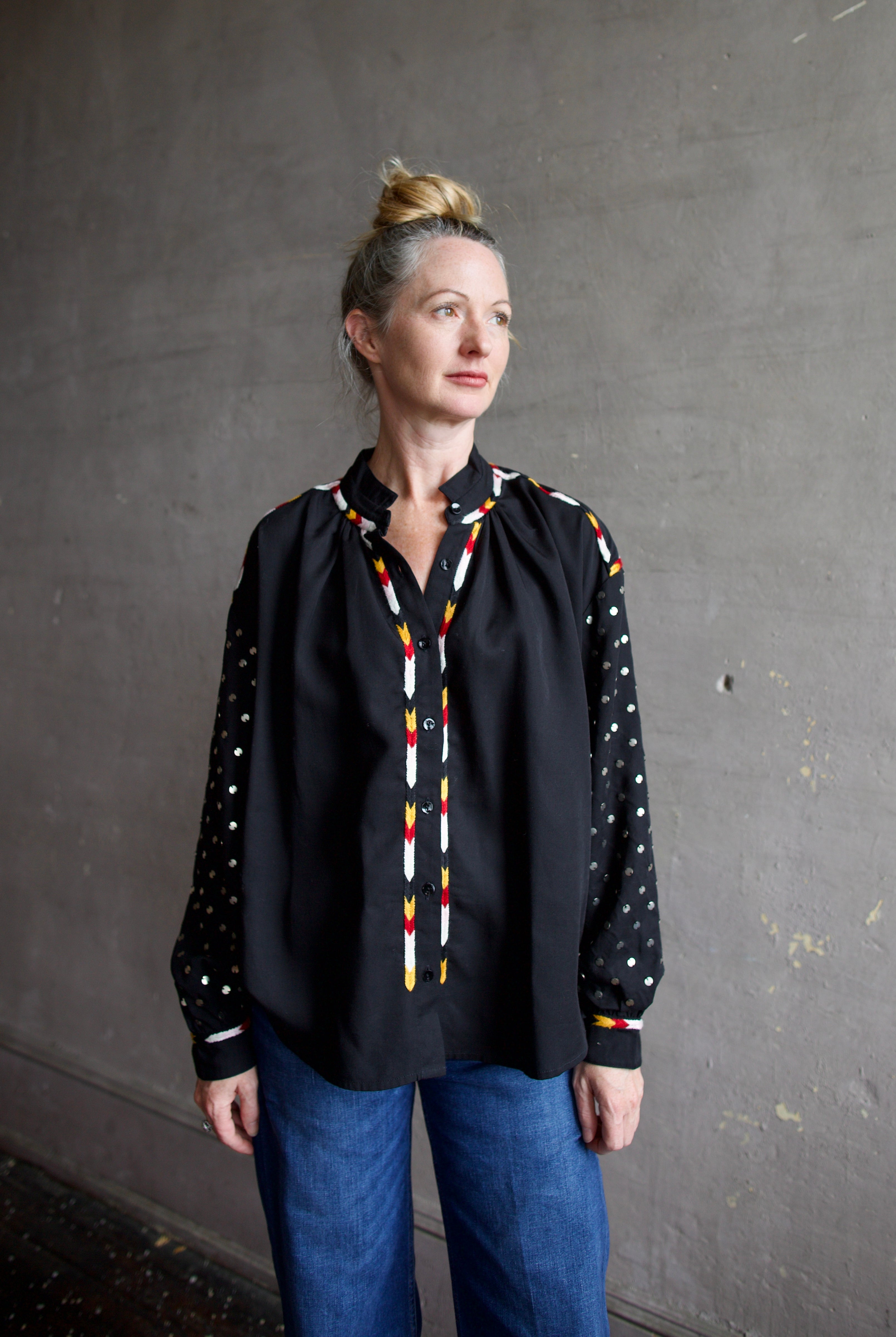 Image featuring a woman wearing the Alix of Bohemia Kiki Stardust button front black shirt with red, white, yellow, & black embroidery and metal discs on the long sleeves.