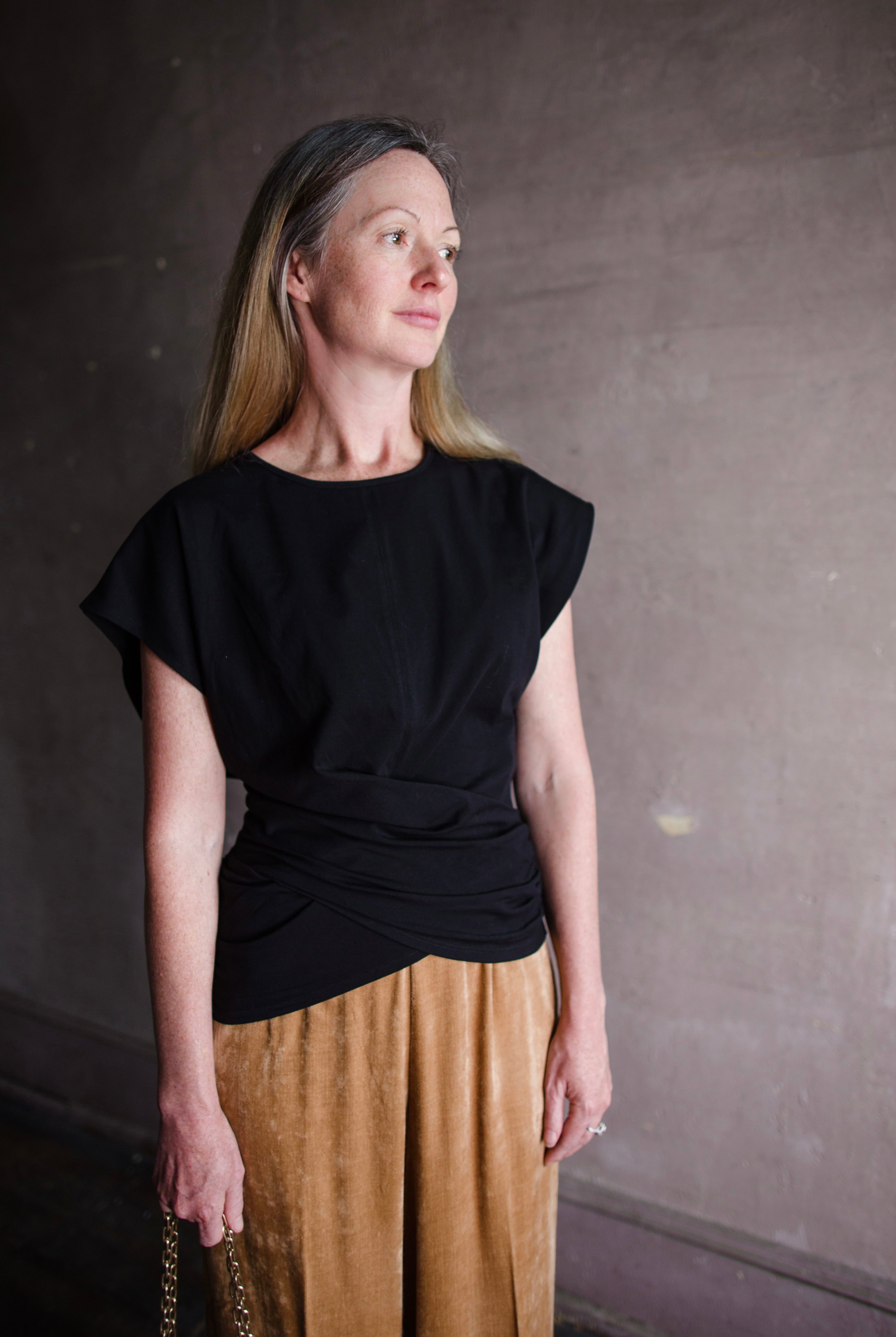 Image featuring a woman wearing the twist top by Apiece Apart with a crew neckline,  cap sleeve and a flattering twist at the waist in black.