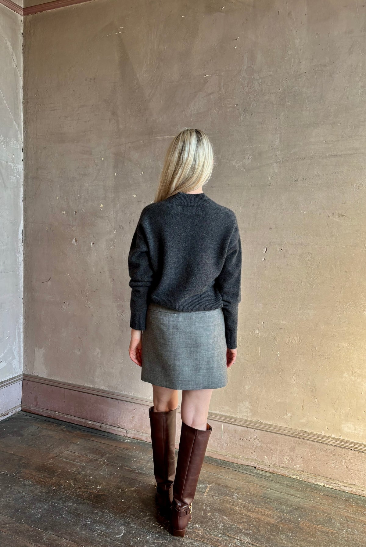 Mature woman stands with her back to the camera wearing a gray textured Marta mini skirt from Apiece Apart with brown knee-high leather boots perfect for Fall