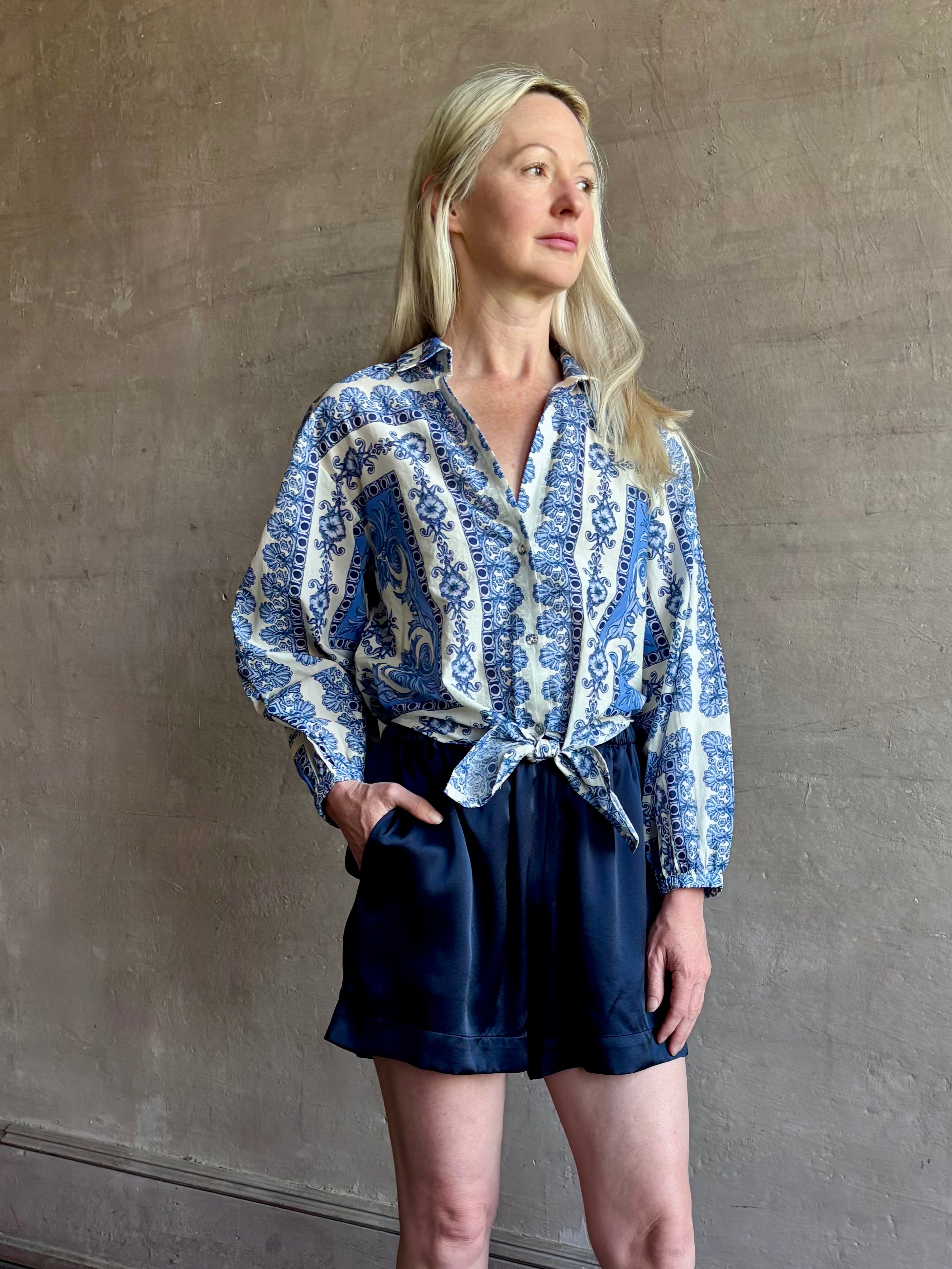 Mature woman wearing the Rumson Top from Cara Cara rendered in a blue and white Hudson Scarf Sky print and crafted from 100% cotton with long sleeves, button down v- neck and tie front cropped top