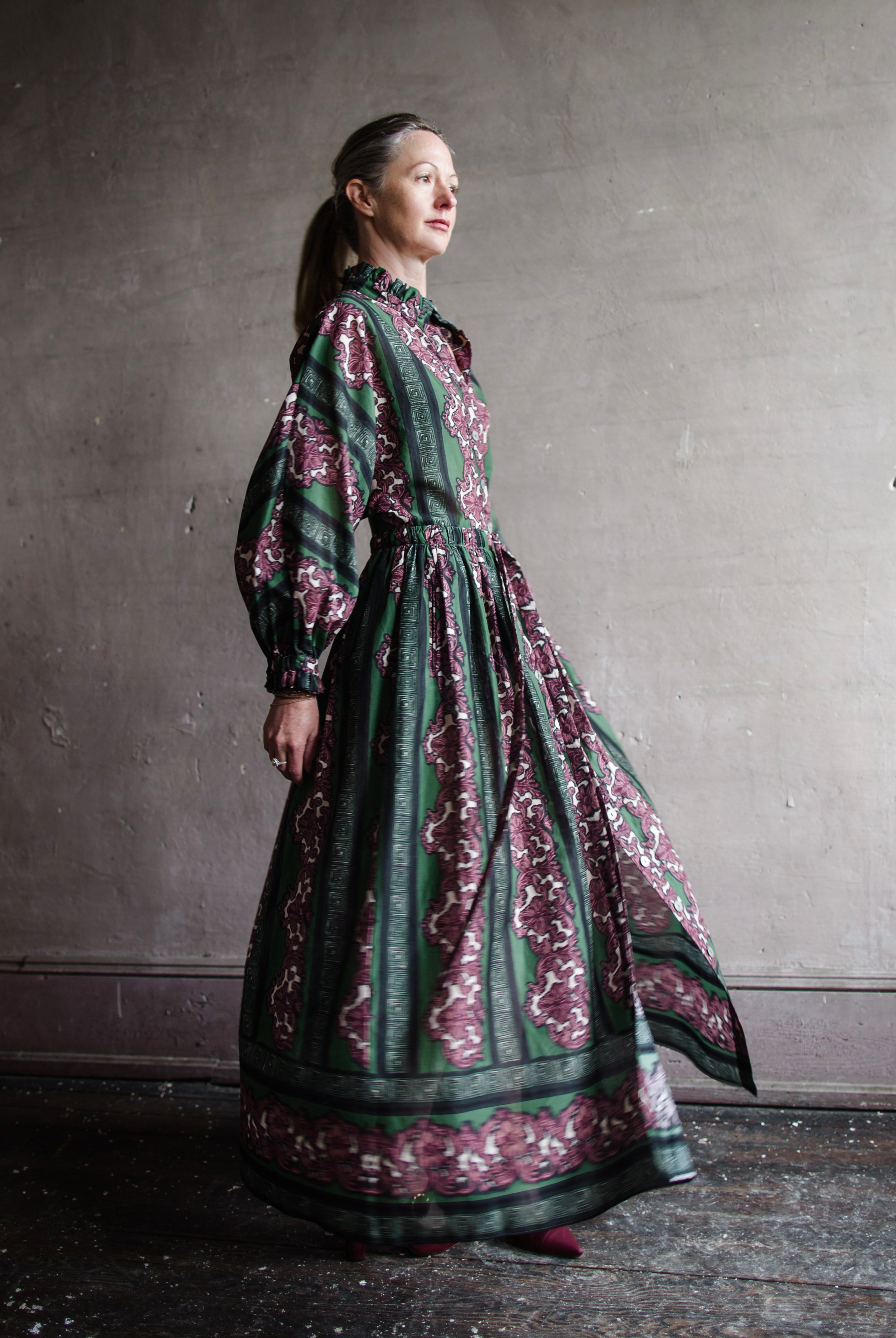 Image features woman wearing The Beatrice dress by Cara Cara. Dress has a high neckline with a button front and drawstring waist with Eme Mauve Damask print.