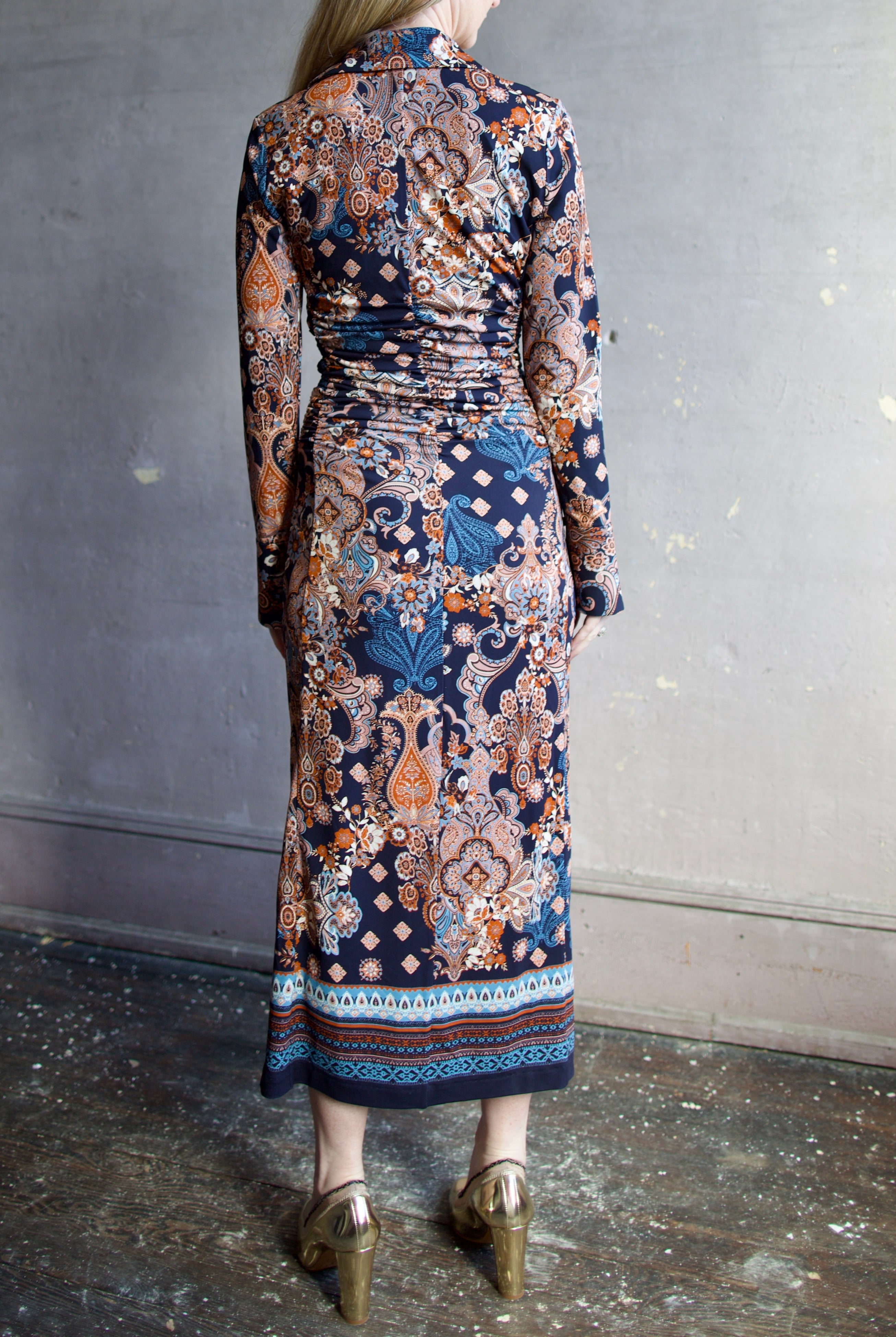 Image featuring a woman wearing the Shirley dress by Cara Cara with a button front, ruched body and side slits in a tailored fit paisley print in blues, orange, and cream.