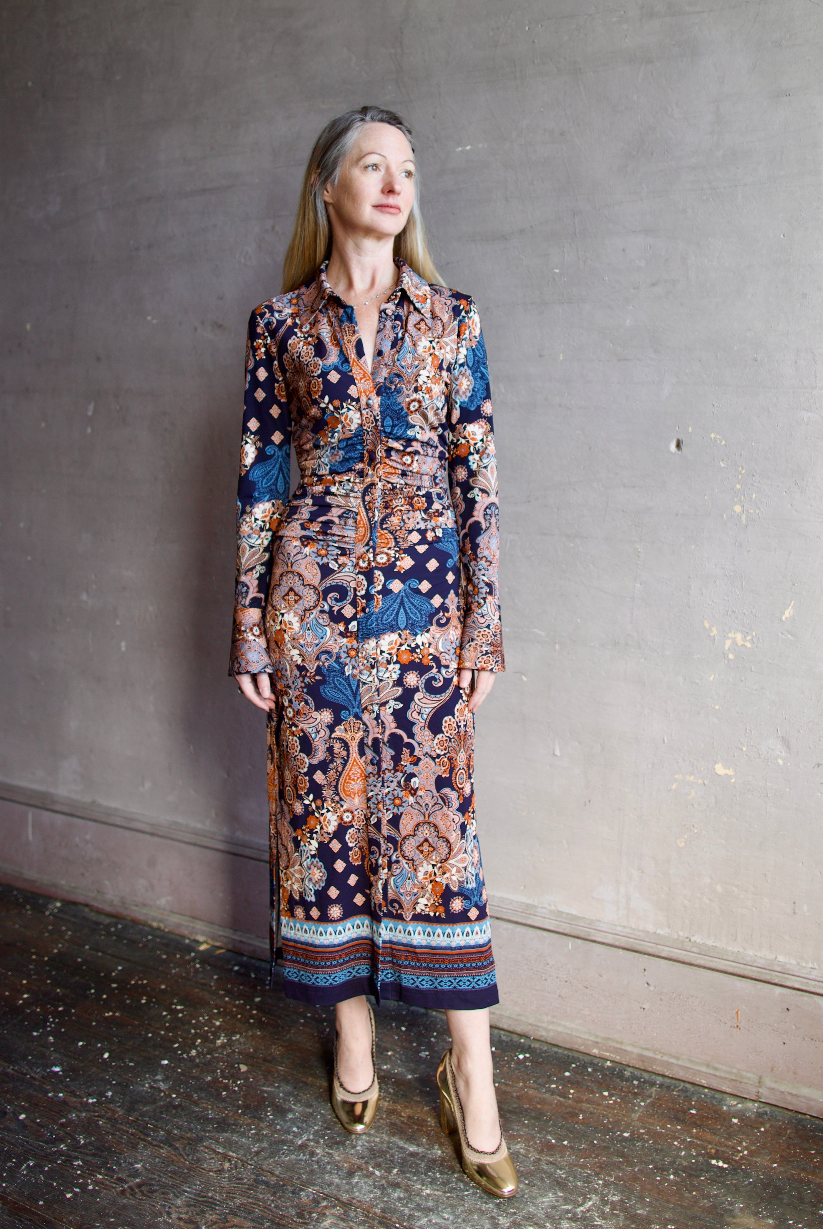Image featuring a woman wearing the Shirley dress by Cara Cara with a button front, ruched body and side slits in a tailored fit paisley print in blues, orange, and cream.