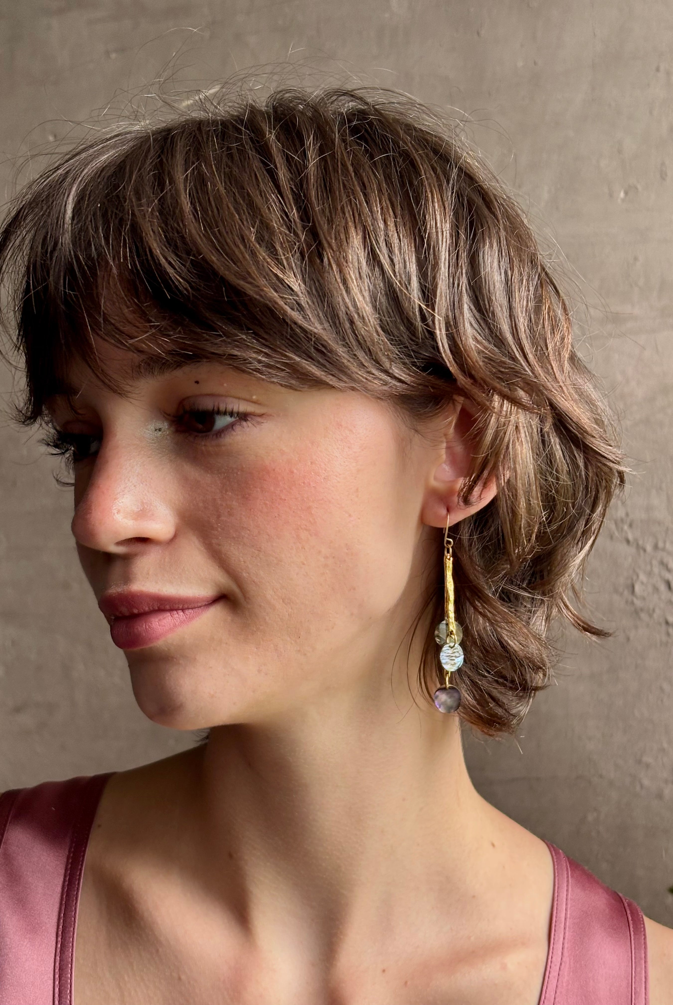 Woman wearing Lyra Earrings from designer jewelry artist Emilie Shapiro with iridescent paua shells dangling from textured brass and a gold filled wire hook with beautiful movement handcrafted in USA