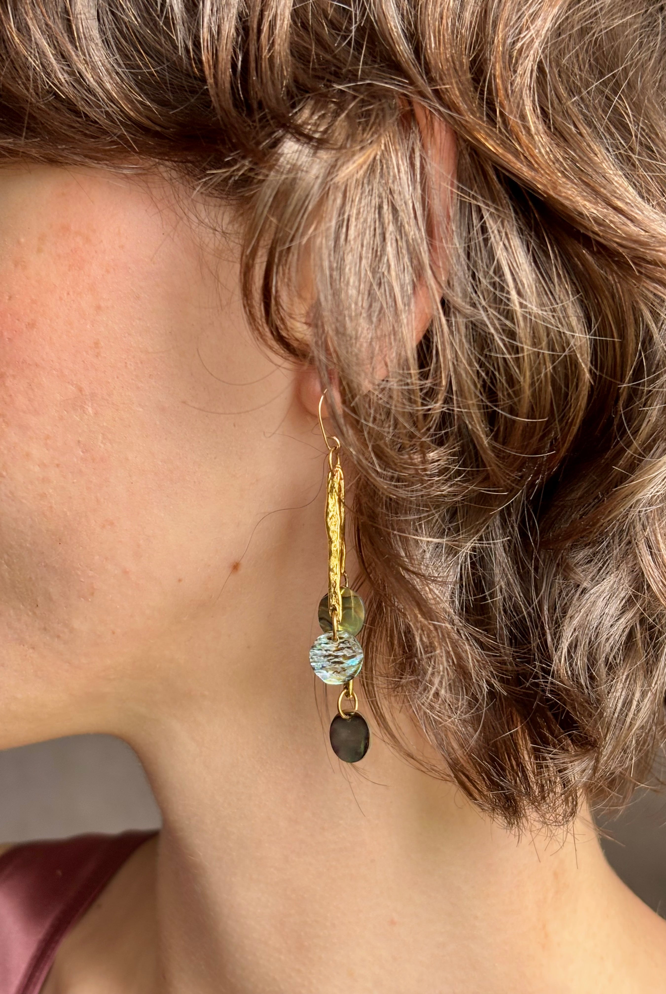 Woman wearing Lyra Earrings from designer jewelry artist Emilie Shapiro with iridescent paua shells dangling from textured brass and a gold filled wire hook with beautiful movement handcrafted in USA