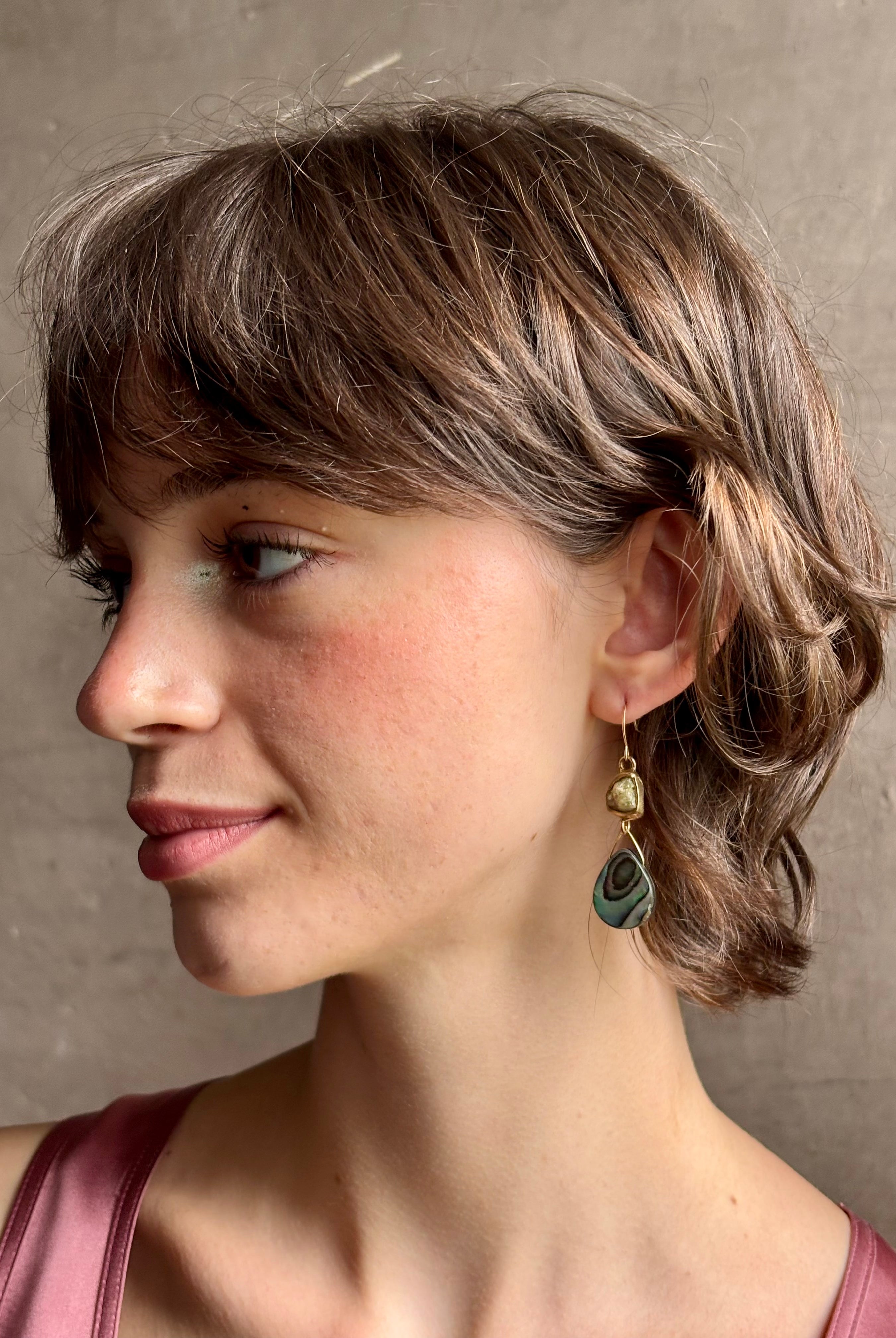 Woman wearing Northern Light Earrings from jewelry artist Emilie Shapiro with a tear shaped paua shell dangling from a natural garnet mounted in brass and suspended from a gold filled hook handcrafted in USA