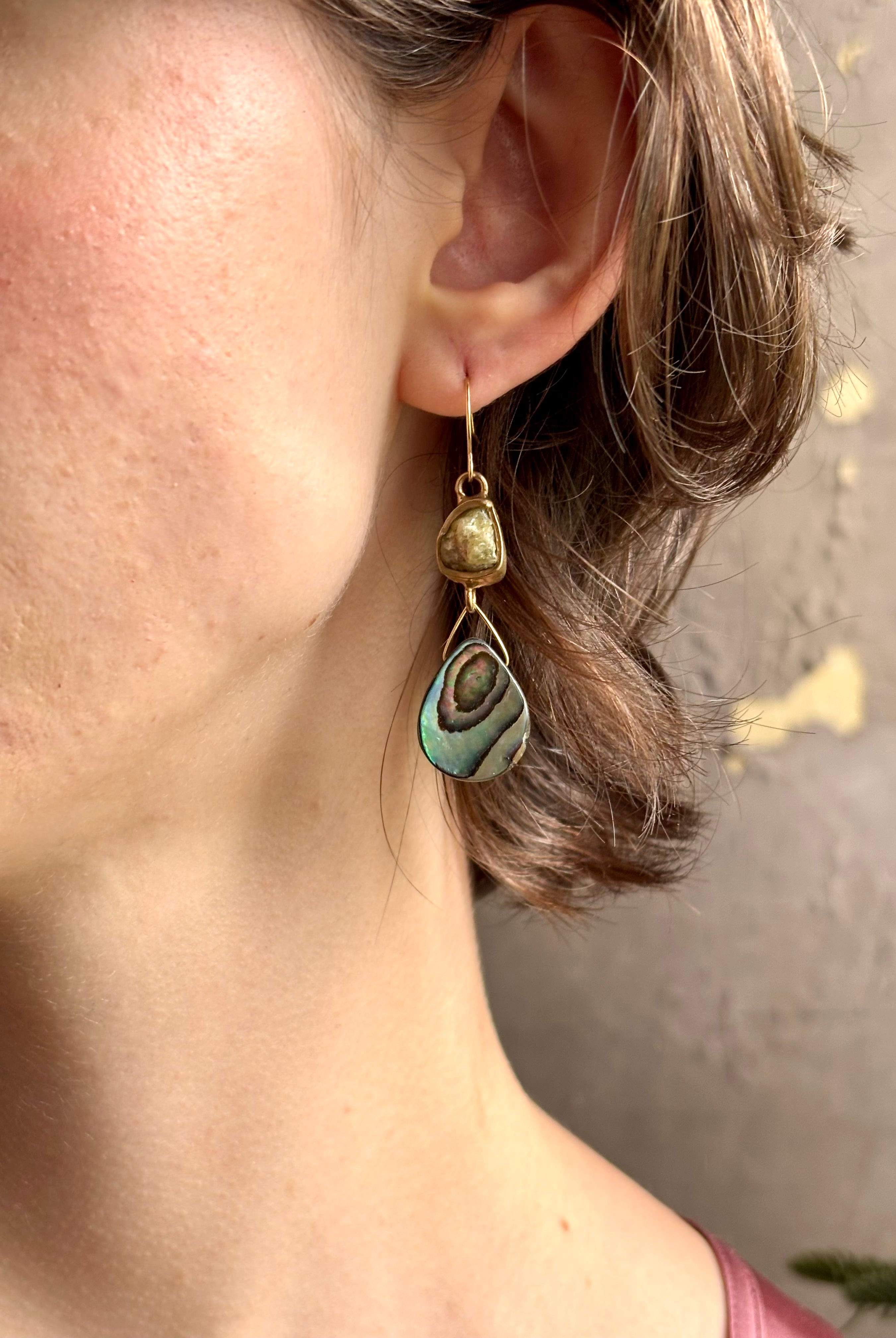 Woman wearing Northern Light Earrings from jewelry artist Emilie Shapiro with a tear shaped paua shell dangling from a natural garnet mounted in brass and suspended from a gold filled hook handcrafted in USA