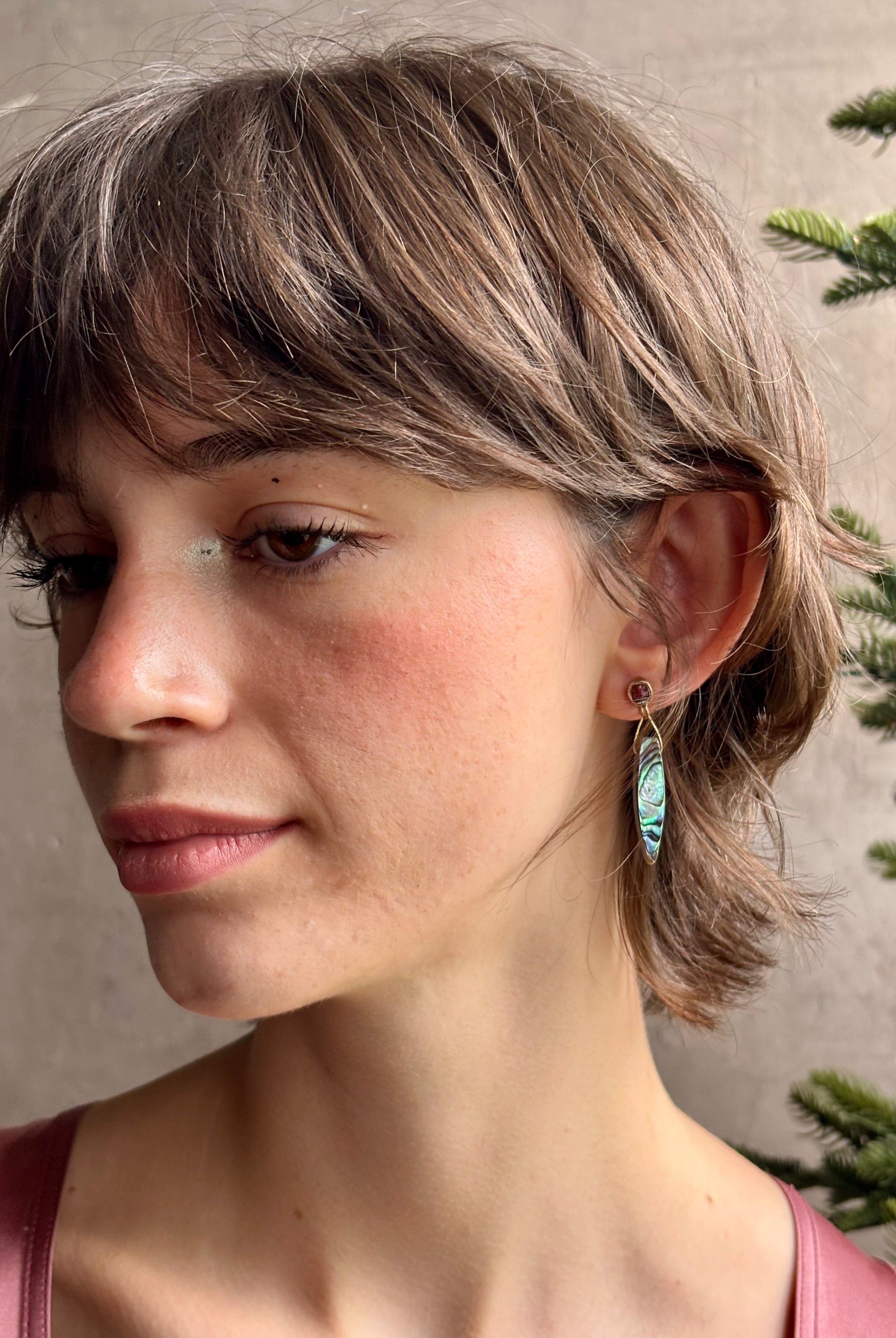 Woman wearing Umbra Earrings from fine jewelry designer Emilie Shapiro with a delicate pink spinel stud anchoring an oval paua shell dandling from a brass triangle wire made in USA