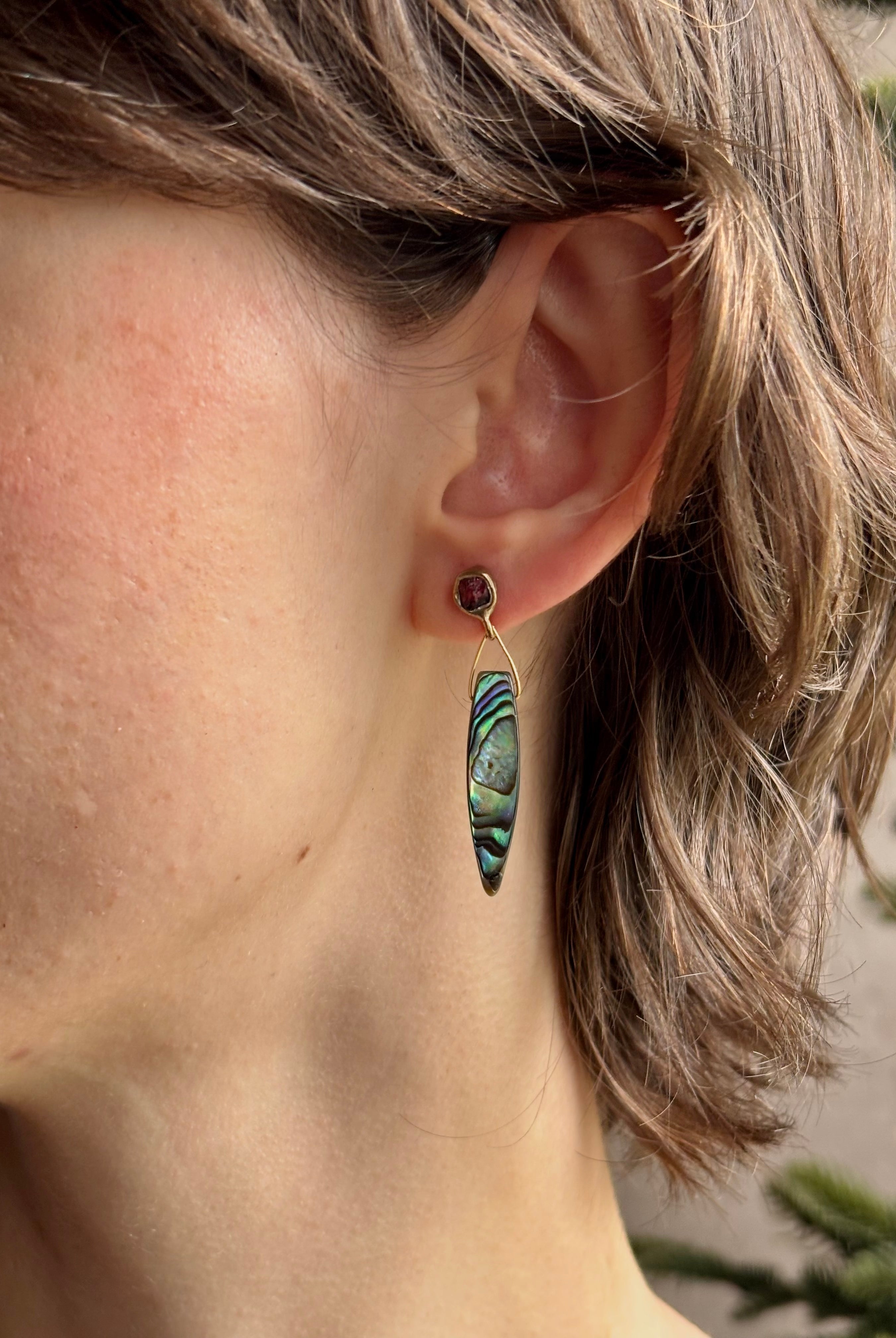 Woman wearing Umbra Earrings from fine jewelry designer Emilie Shapiro with a delicate pink spinel stud anchoring an oval paua shell dandling from a brass triangle wire made in USA