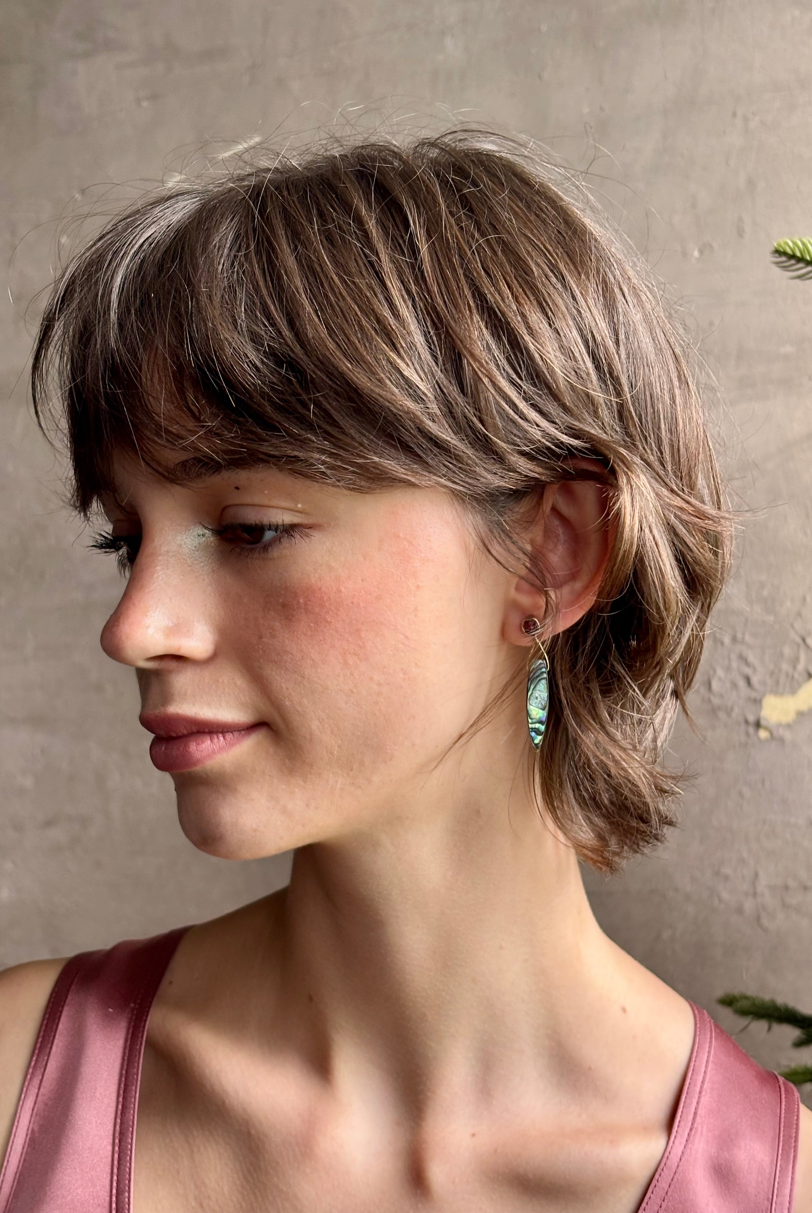 Woman wearing Umbra Earrings from fine jewelry designer Emilie Shapiro with a delicate pink spinel stud anchoring an oval paua shell dandling from a brass triangle wire made in USA