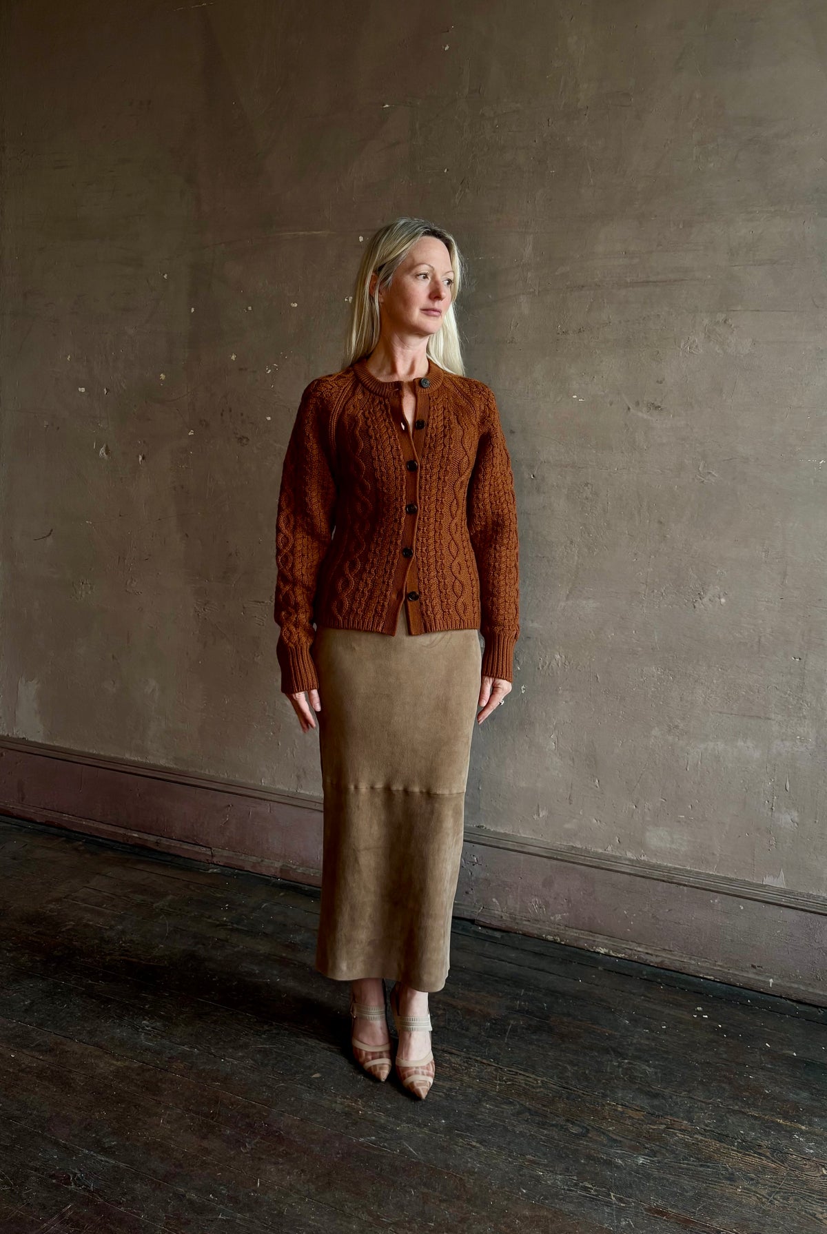 Mature woman wearing Guest in Residence's Cable Sculpted Cardigan Burnt Sienna wool-cashmere cable knit raglan sleeves. Richly textured heirloom-quality warmth