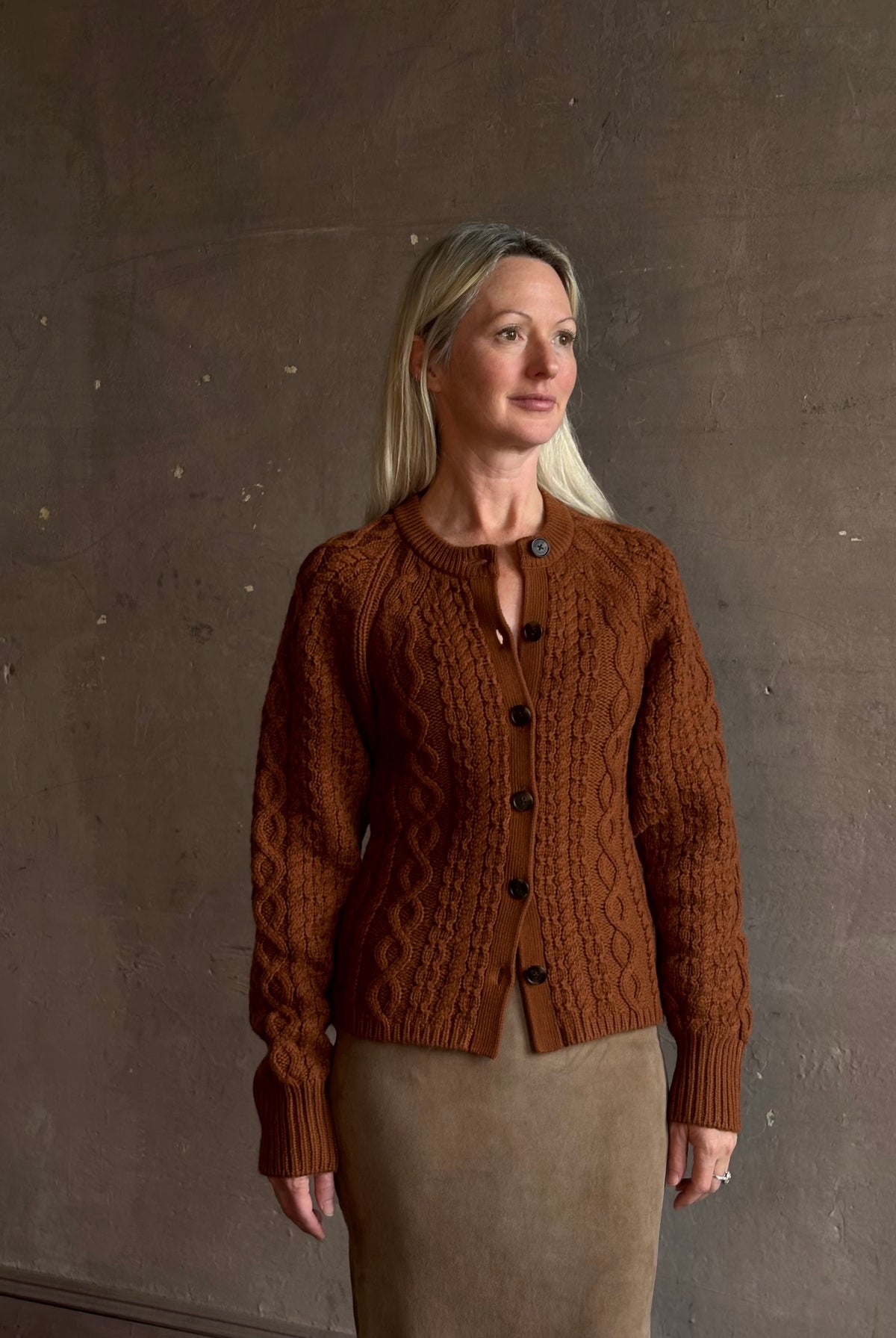 Mature woman wearing Guest in Residence's Cable Sculpted Cardigan Burnt Sienna wool-cashmere cable knit raglan sleeves. Richly textured heirloom-quality warmth
