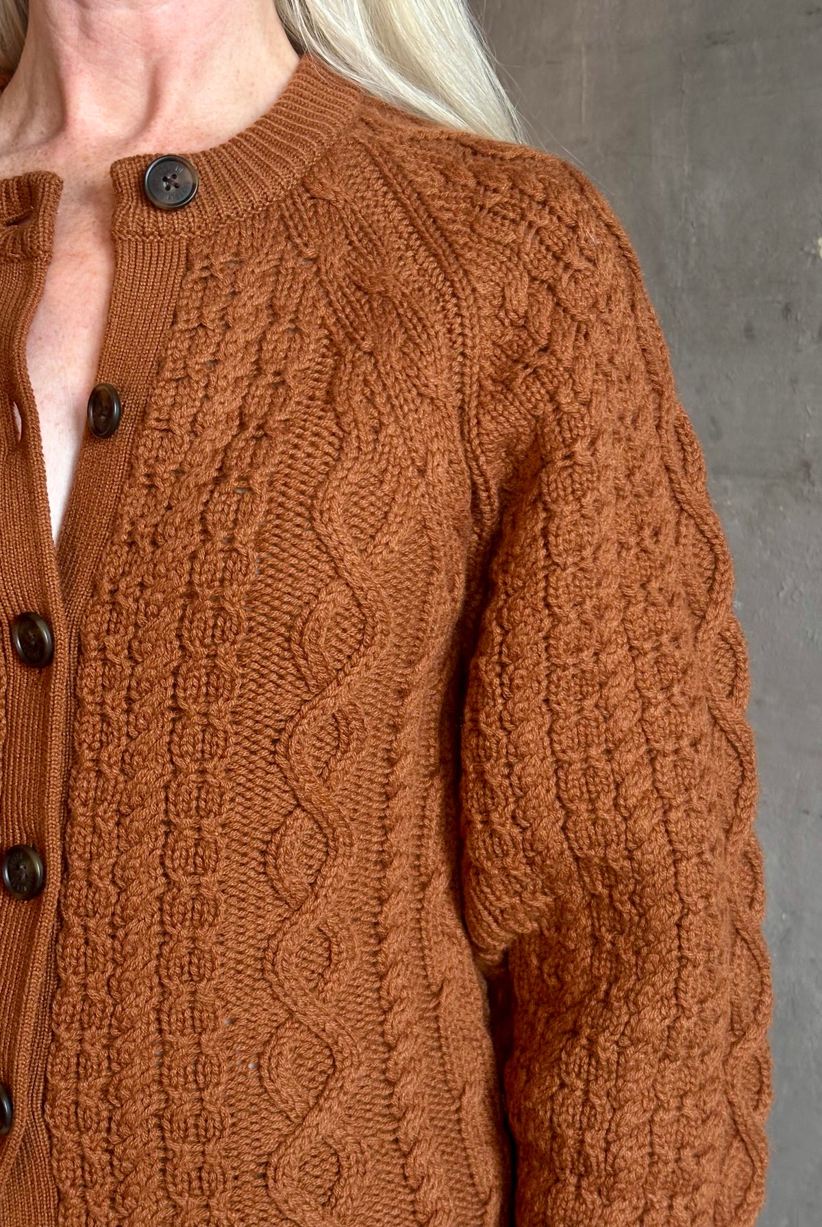 Mature woman wearing Guest in Residence's Cable Sculpted Cardigan Burnt Sienna wool-cashmere cable knit raglan sleeves. Richly textured heirloom-quality warmth