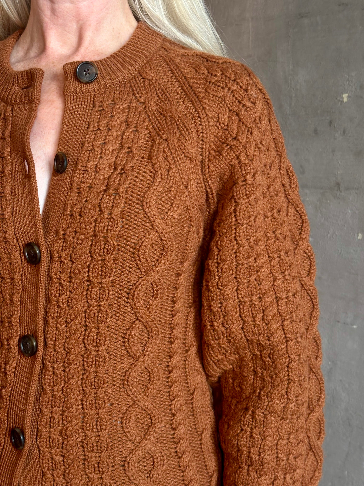 Mature woman wearing Guest in Residence's Cable Sculpted Cardigan Burnt Sienna wool-cashmere cable knit raglan sleeves. Richly textured heirloom-quality warmth