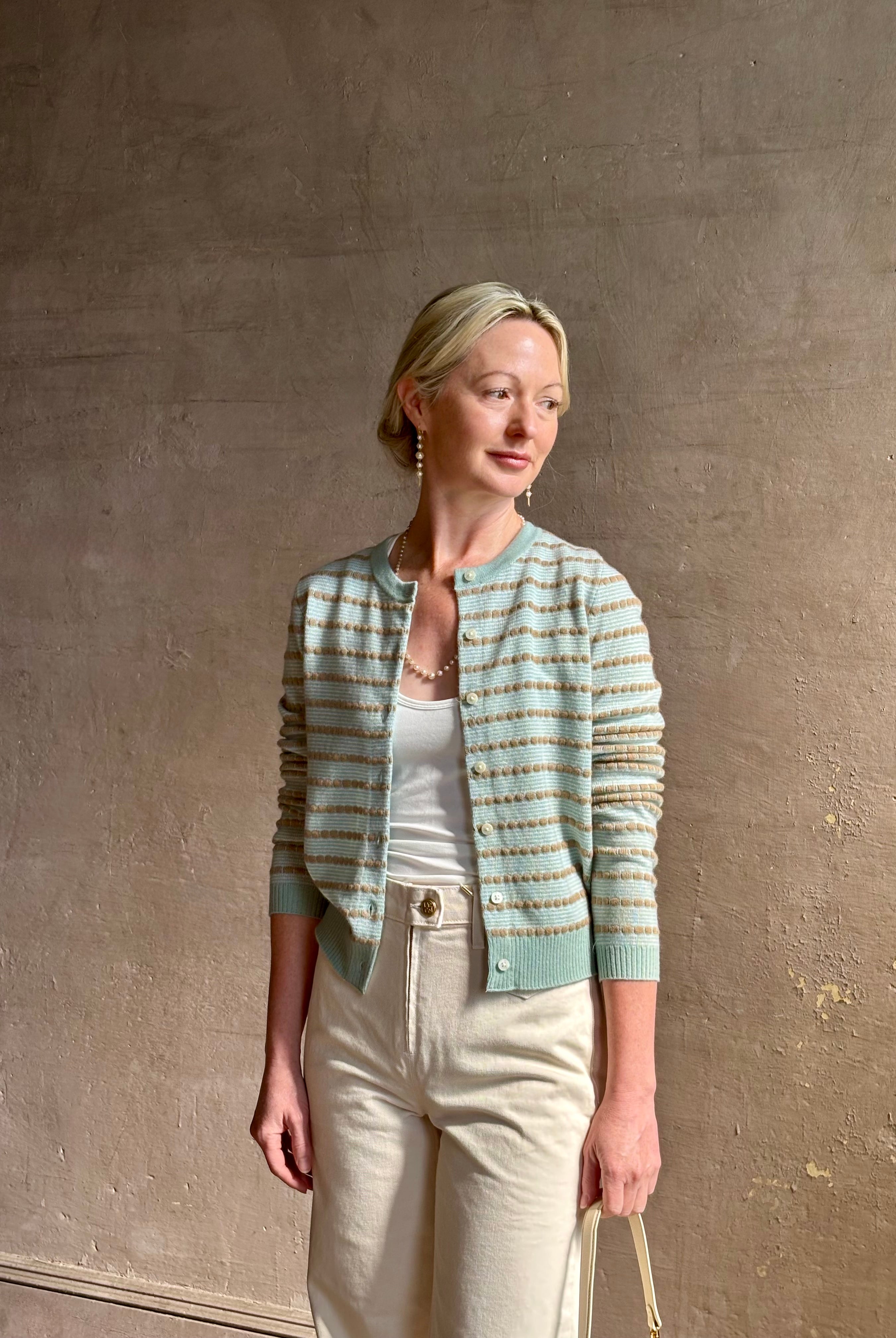 Mature woman wearing Pebble Stripe Jane Cardigan from Guest in Residence rendered in a serene sage brush aqua Khaki combo and crafted from 100% cashmere with crew neck, long sleeves and button front