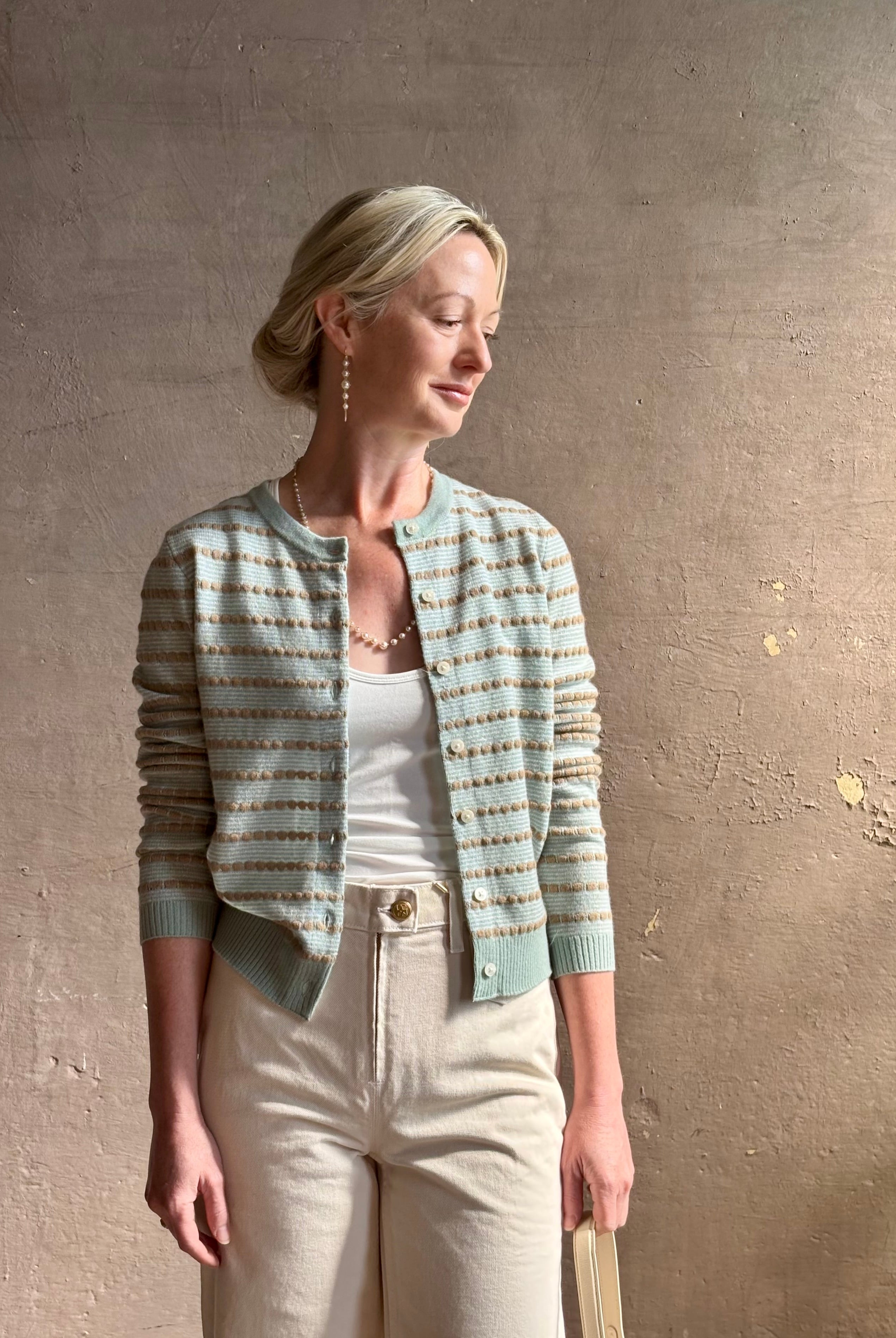 Mature woman wearing Pebble Stripe Jane Cardigan from Guest in Residence rendered in a serene sage brush aqua Khaki combo and crafted from 100% cashmere with crew neck, long sleeves and button front