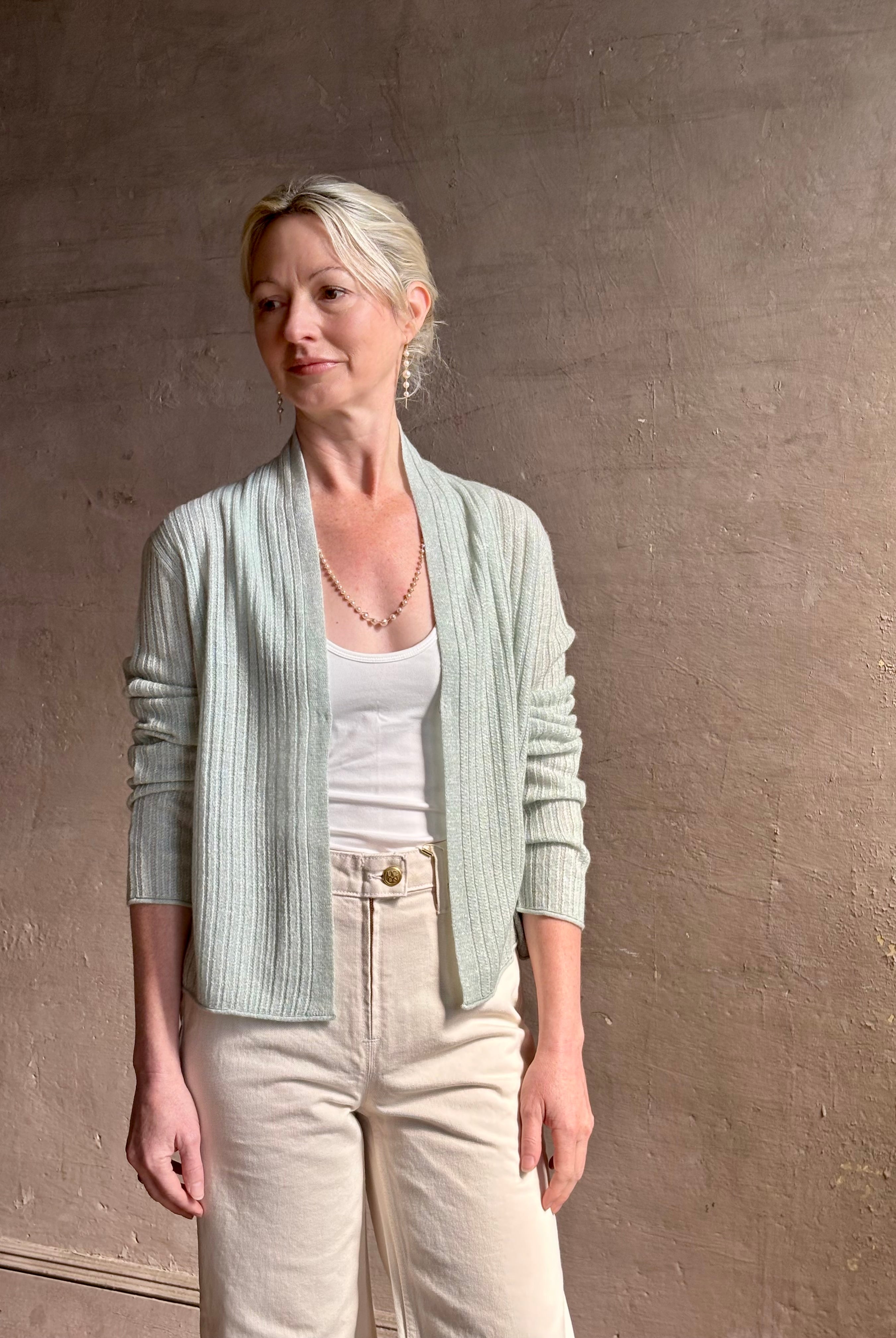 Mature woman wearing Rib Stealth Cardigan from Guest in Residence rendered in a serene sage brush aqua green and crafted from a refined blend of 58% cashmere and 42% linen with long sleeves and a hidden hook and eye closure