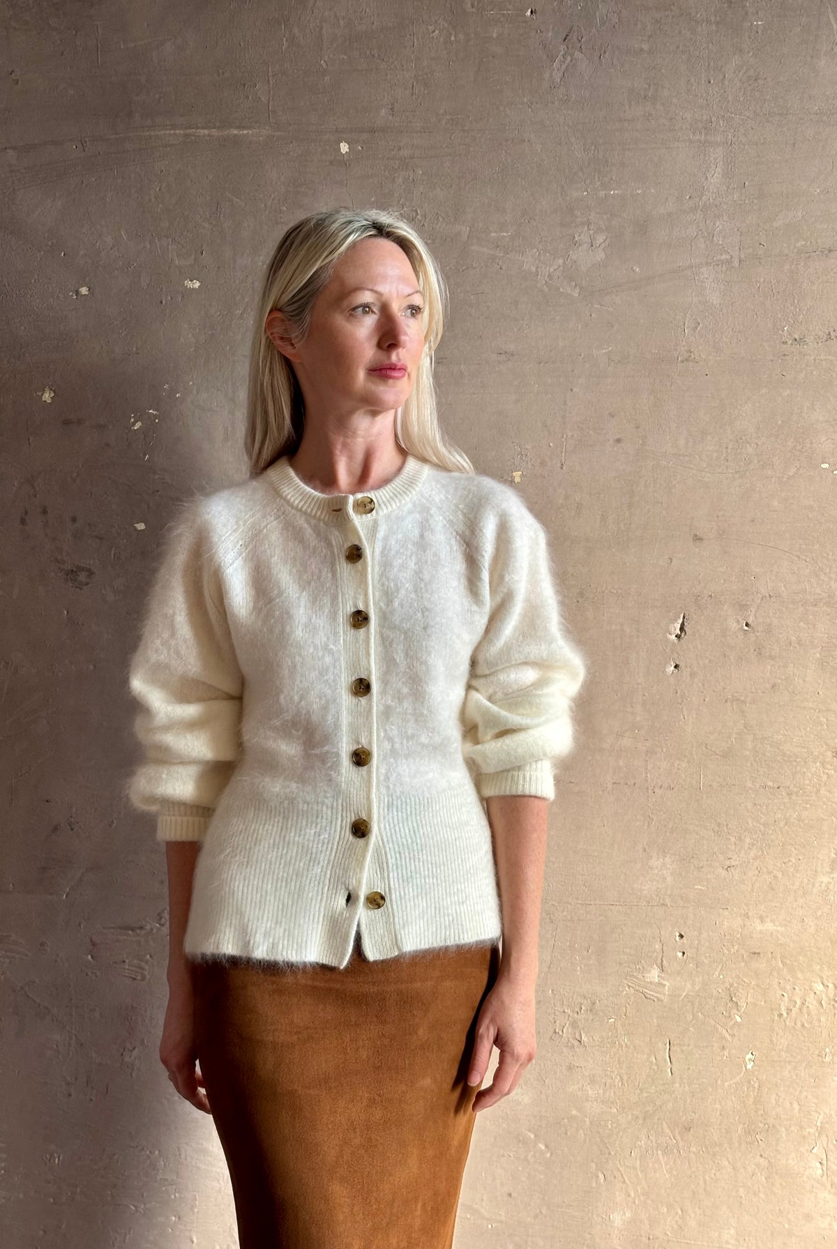 A woman wearing a cream-colored fuzzy knit cardigan with brown buttons and a brown skirt, standing against a textured beige wall.