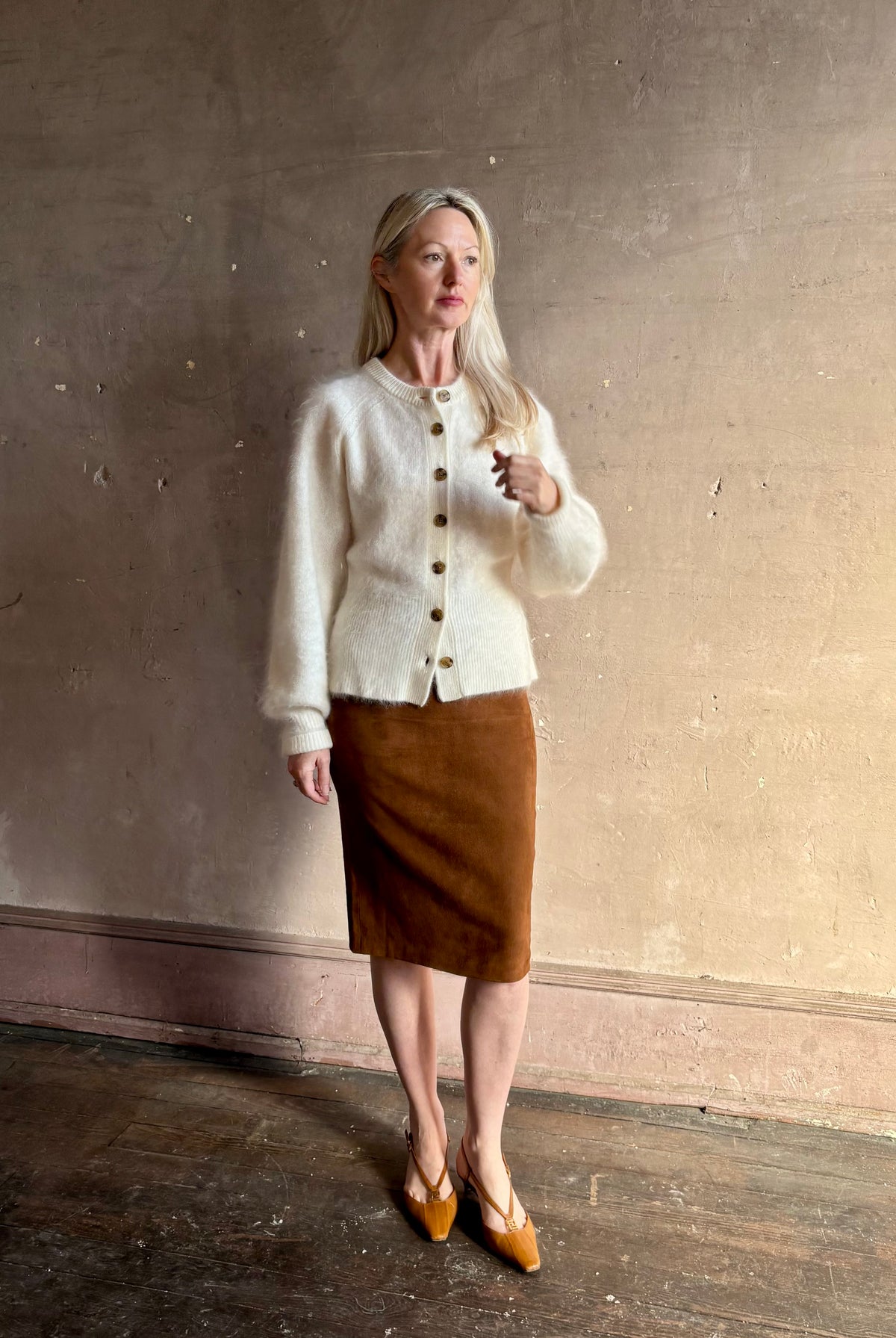 A woman stands against a textured beige wall wearing a cream-colored fuzzy button-front cardigan, a brown knee-length skirt, and Fendi Shoes