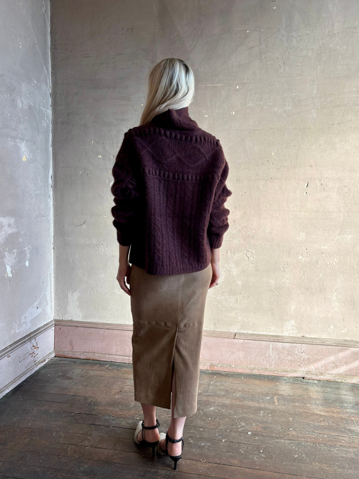Back image of a woman wearing a dark plum purple cable-knit sweater with intricate pattern all the way around the Guest in Residence turtle neck sweater