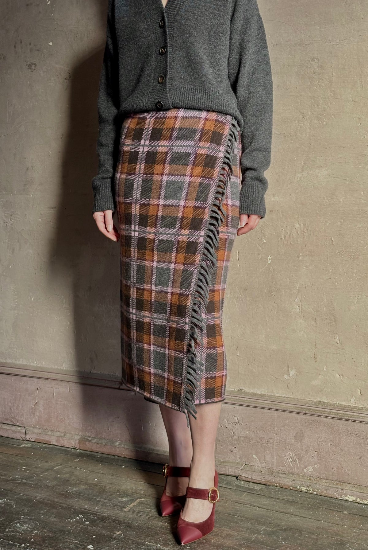 Image of mature woman wearing a gray cardigan with the Guest in Residence Tartan wrap pencil skirt in brown, orange, and gray plaid with fringe trim