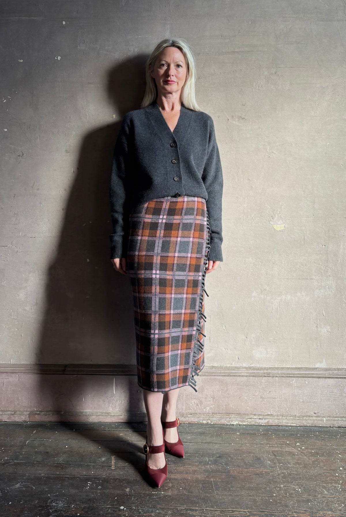 Image of a mature woman wearing a dark gray button-up cardigan styled with Gigi Hadid Guest in Residence grey, brown and orange plaid midi skirt with fringe detailing