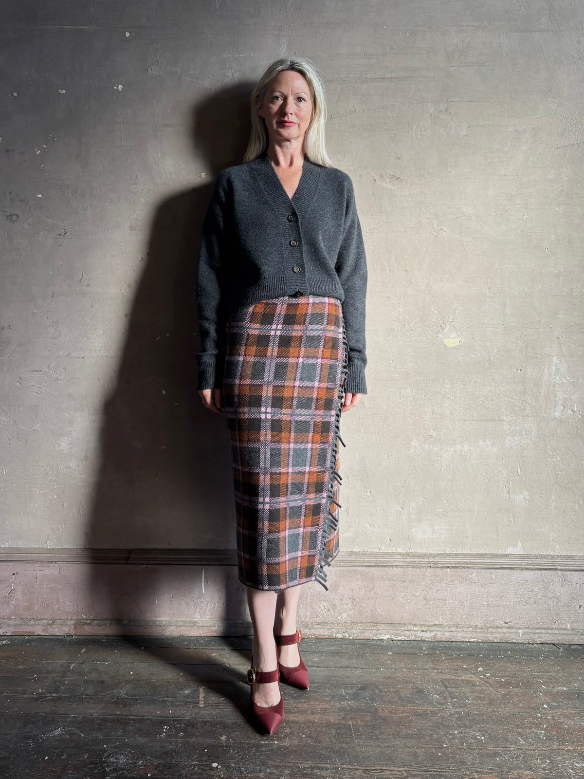 Image of a mature woman wearing a dark gray button-up cardigan styled with Gigi Hadid Guest in Residence grey, brown and orange plaid midi skirt with fringe detailing