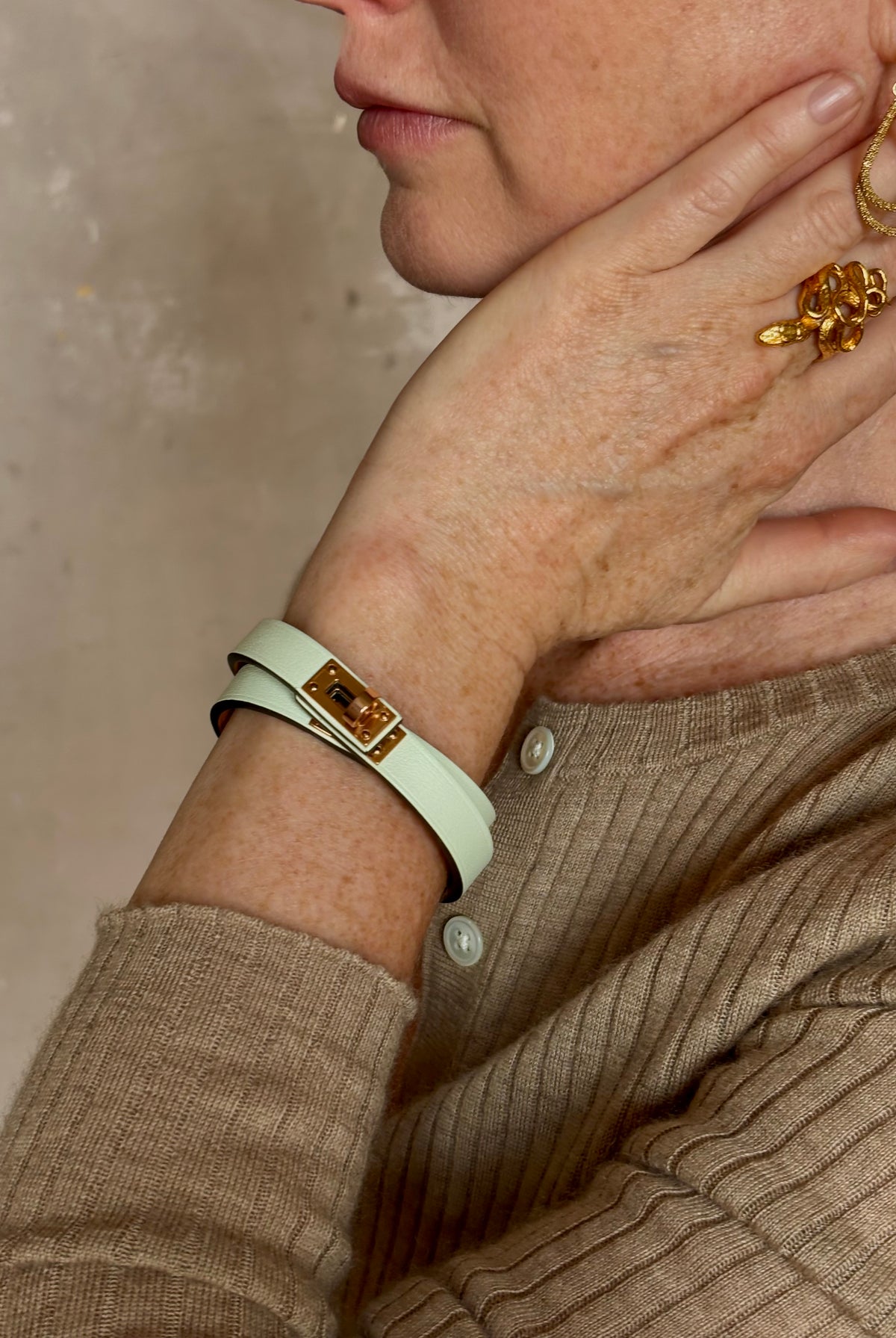 Mature woman wearing Hermès' Mini Kelly Double Tour Wrap Bracelet in Vert d'Eau & Rose Gold pristine, never-worn leather with the iconic Kelly turn-lock. Rare, collectible French luxury