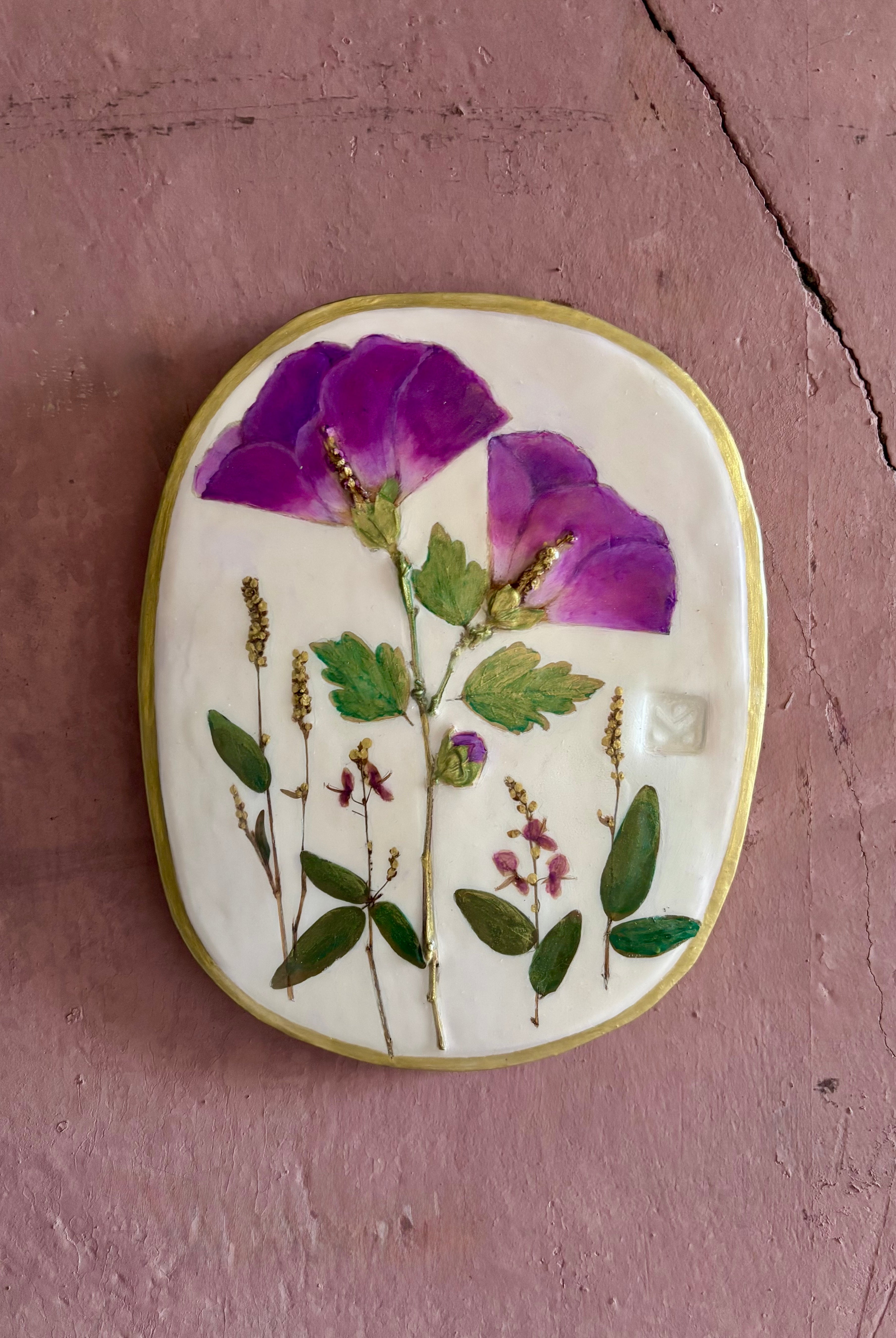 An oval ceramic wall plaque featuring pressed purple rose of sharon hibiscus flowers and green leaves on white clay