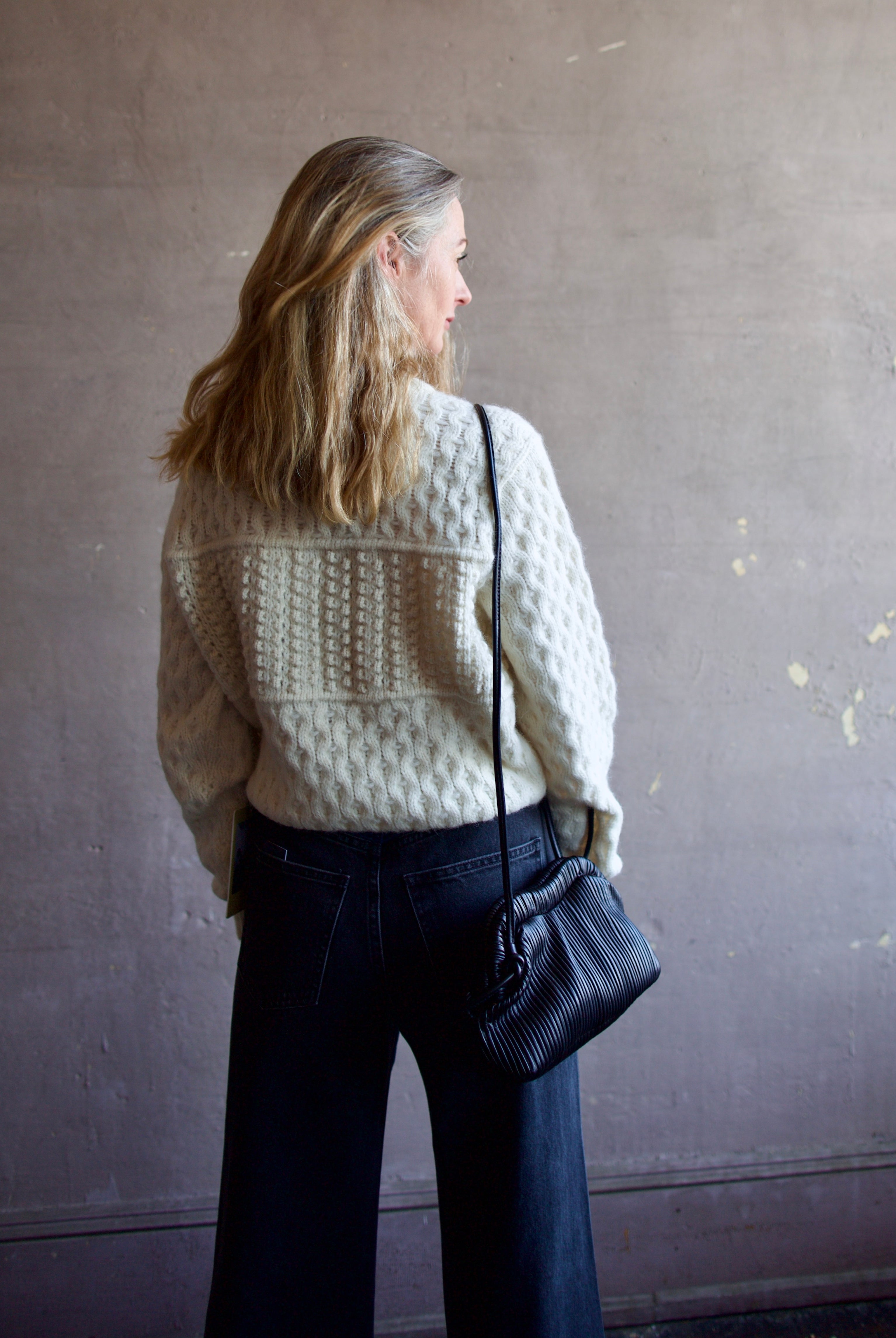 Image featuring a woman wearing the Baci handbag by Mansur Gavriel in black with a magnetic closure, adjustable crossbody strap and intricate pintucked leather across entire hand bag.