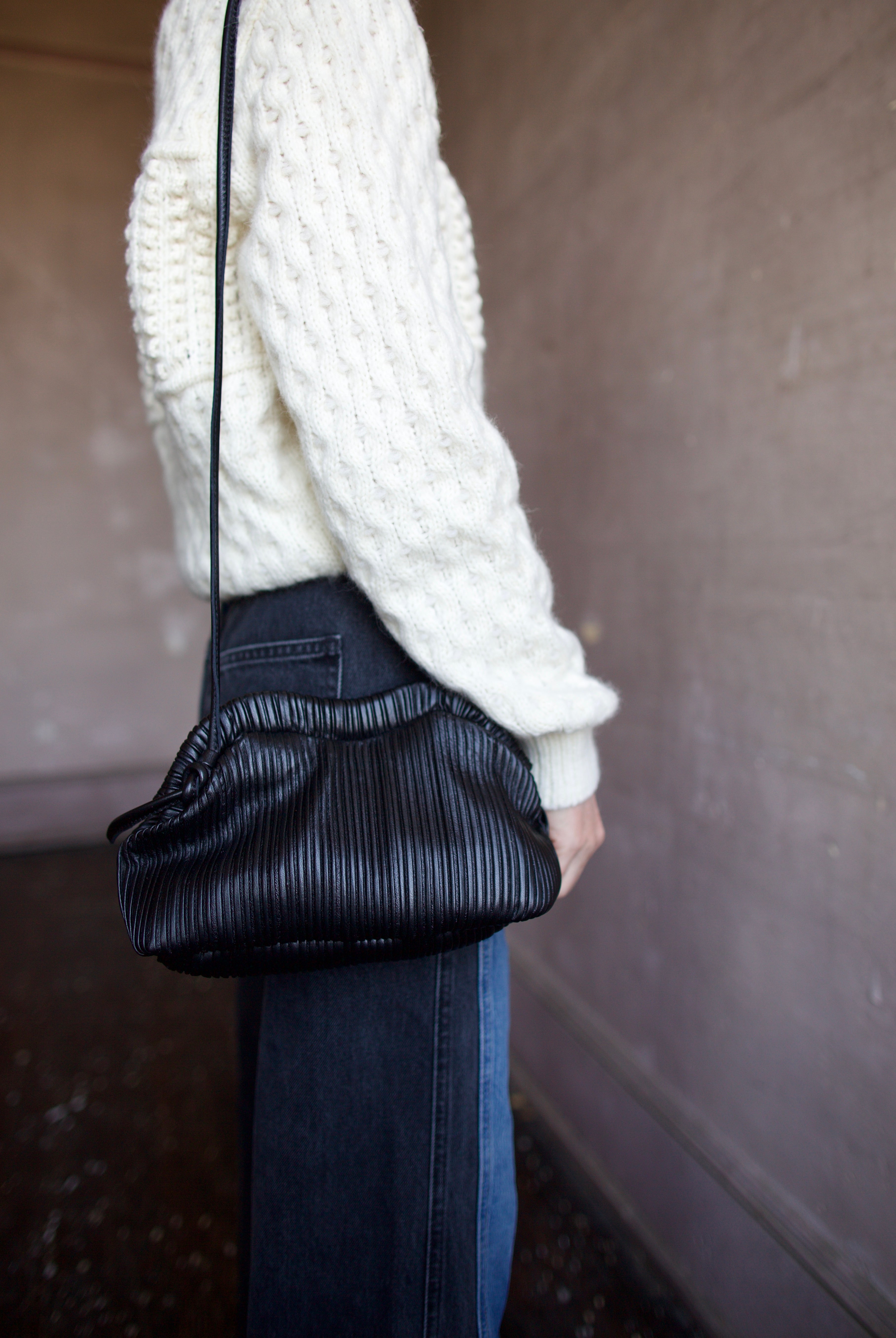 Image featuring a woman wearing the Baci handbag by Mansur Gavriel in black with a magnetic closure, adjustable crossbody strap and intricate pintucked leather across entire hand bag.