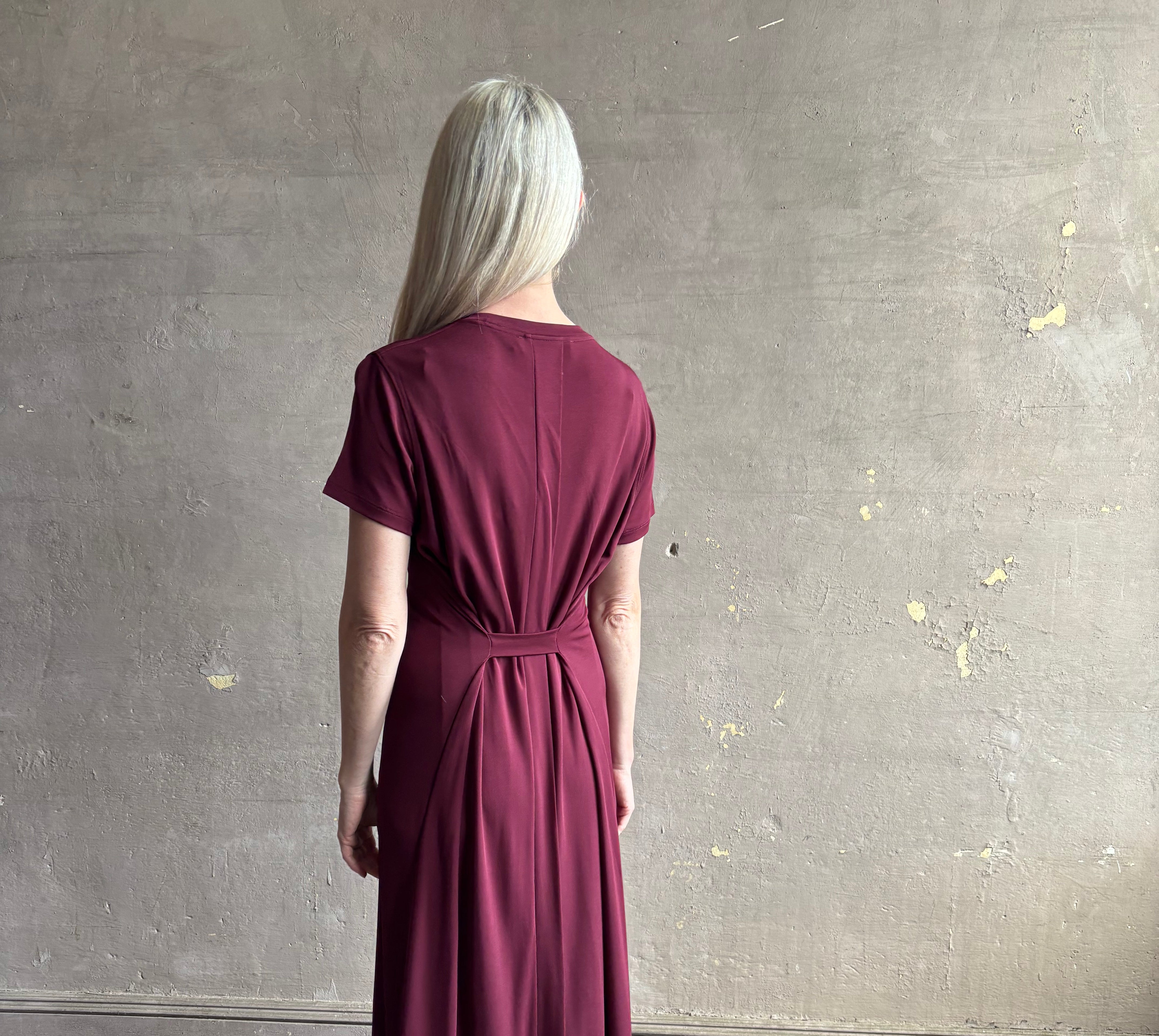 Mature woman wearing the Noelle designer dress from Proenza Schouler in a deep wine hue with elevated back draping detail short sleeves and crew neck