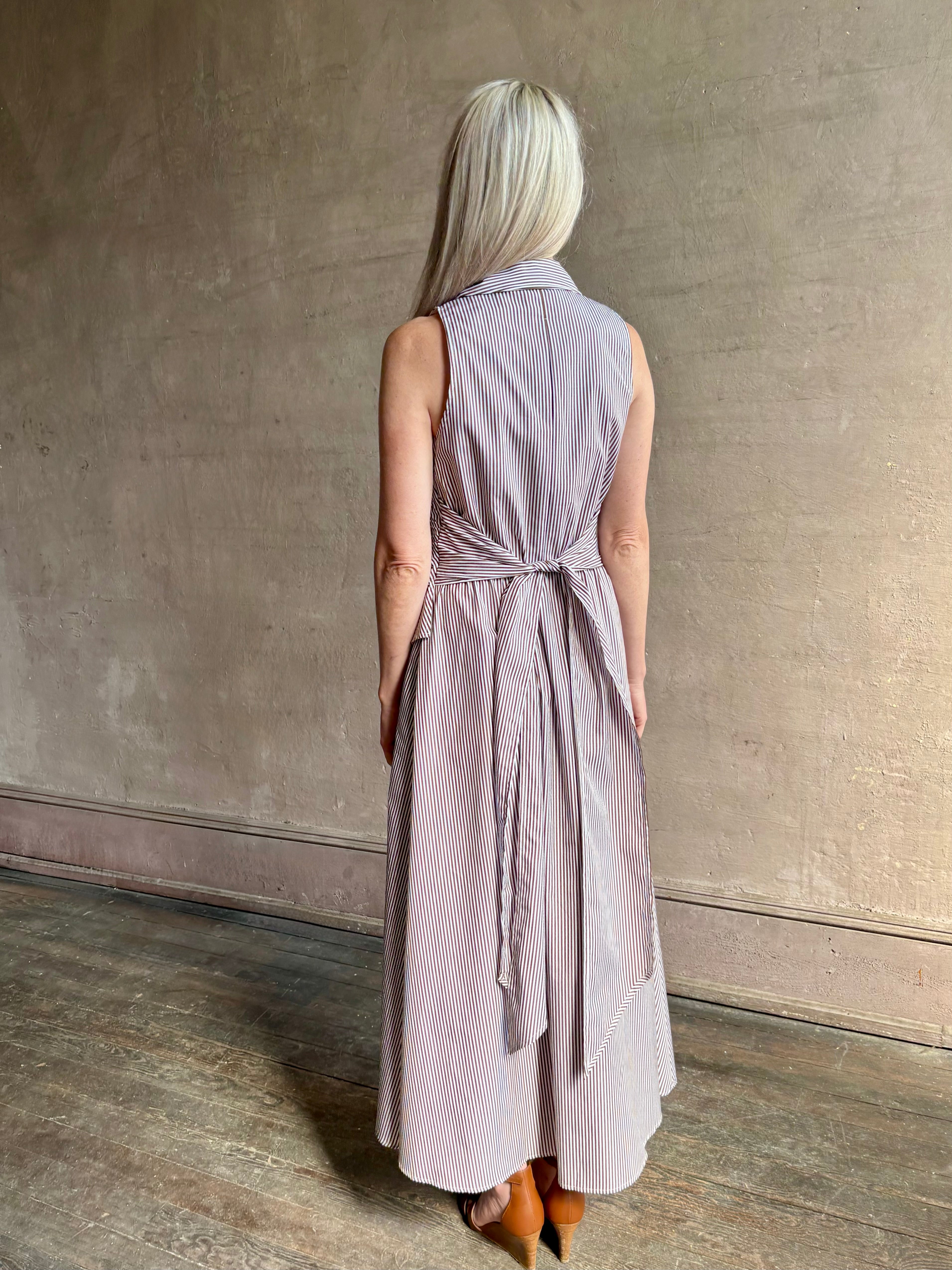 Mature woman wearing the Hannah maxi shirt dress from designer Tanya Taylor rendered in a brown and white stripe and crafted from 100% cotton poplin with a very classy flattering waistline that doesn't stick to body, sleeveless with a collar for a timeless tailored classic dress