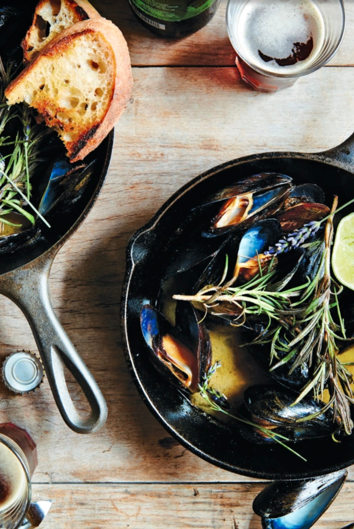 Cast iron skillet with mussels on a rustic wooden table, garnished with rosemary and a lime wedge, alongside toast