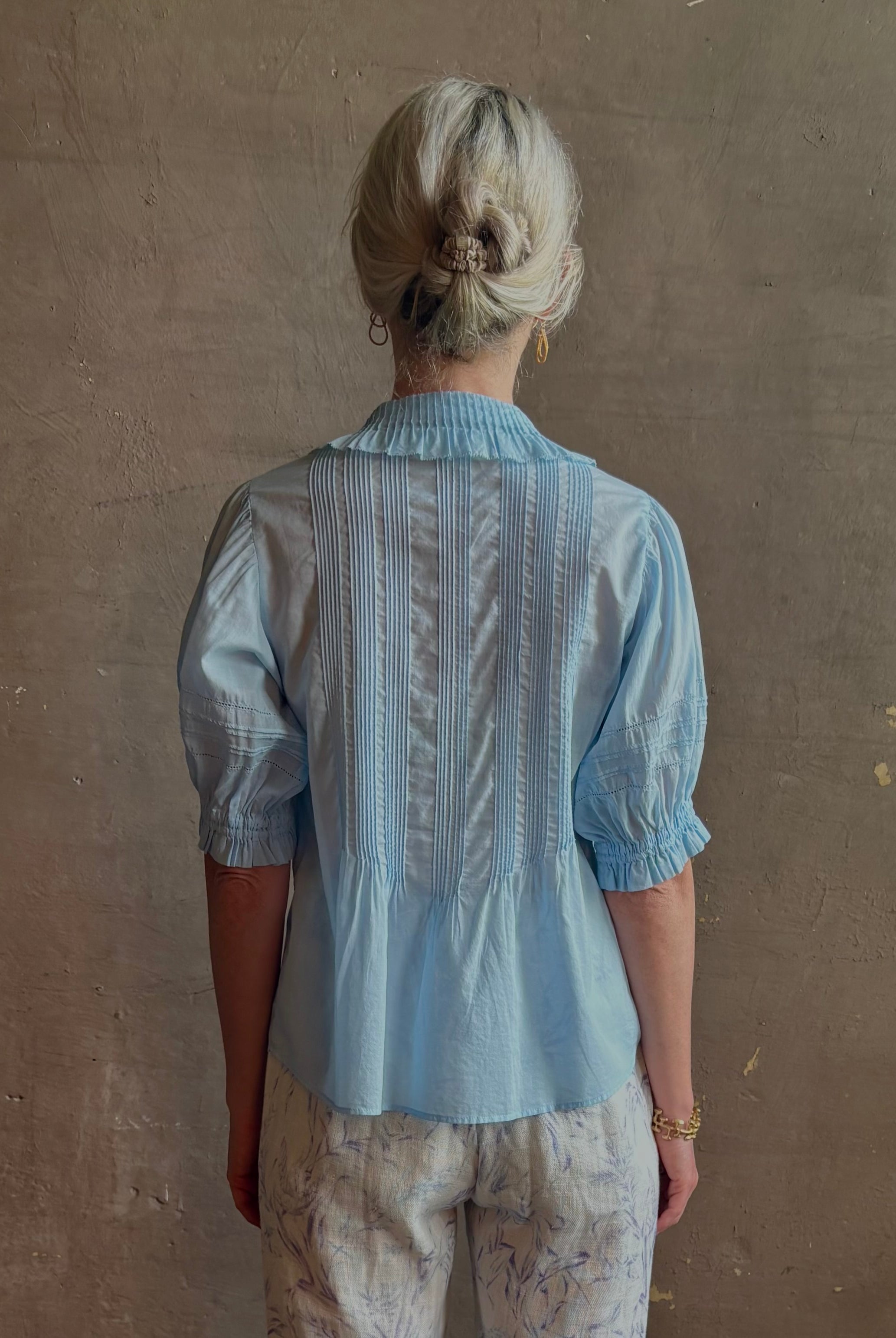 Mature woman wearing Jess Blouse from Trovata rendered in the light blur and crafted from cotton poplin with peter pan collar, half sleeves, intricate pitucking and button front