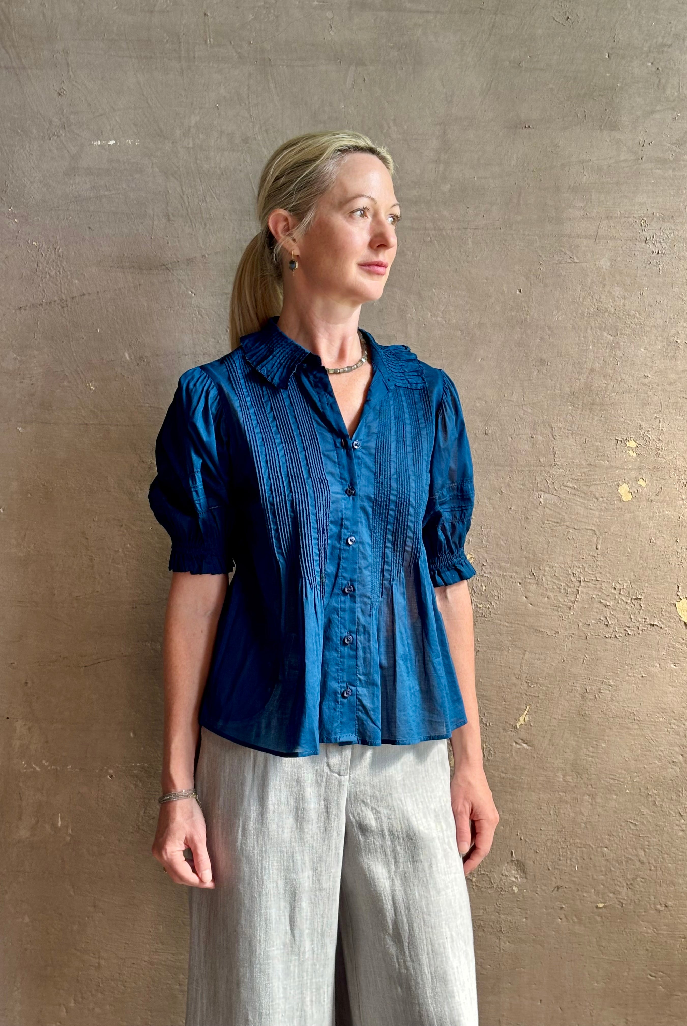 Mature woman wearing Jess Blouse from Trovata rendered in a classic Navy blue and crafted from light weight cotton poplin with short sleeves, peter pan collar, v-neck button front with intricate pintucking details at front and back