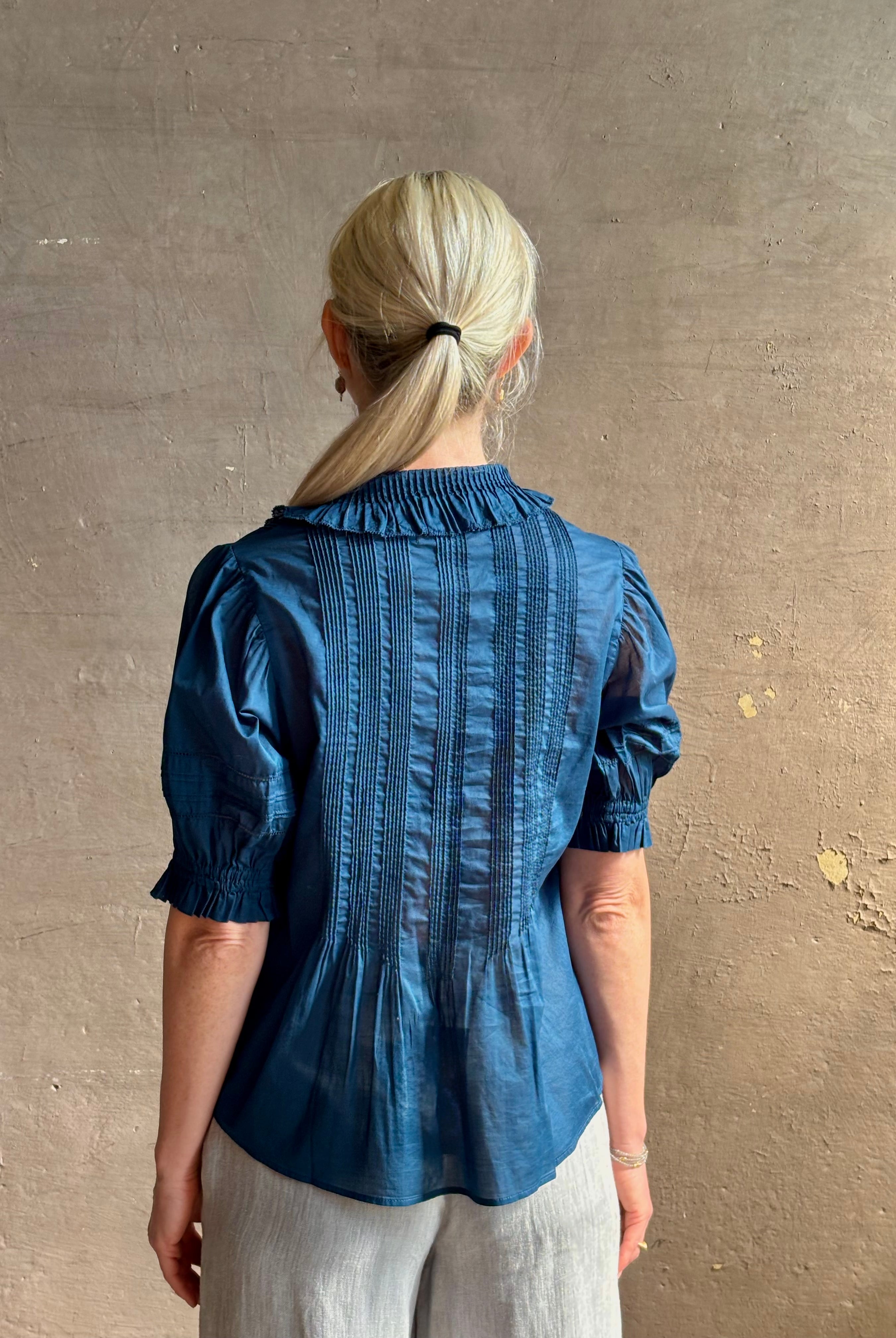 Mature woman wearing Jess Blouse from Trovata rendered in a classic Navy blue and crafted from light weight cotton poplin with short sleeves, peter pan collar, v-neck button front with intricate pintucking details at front and back