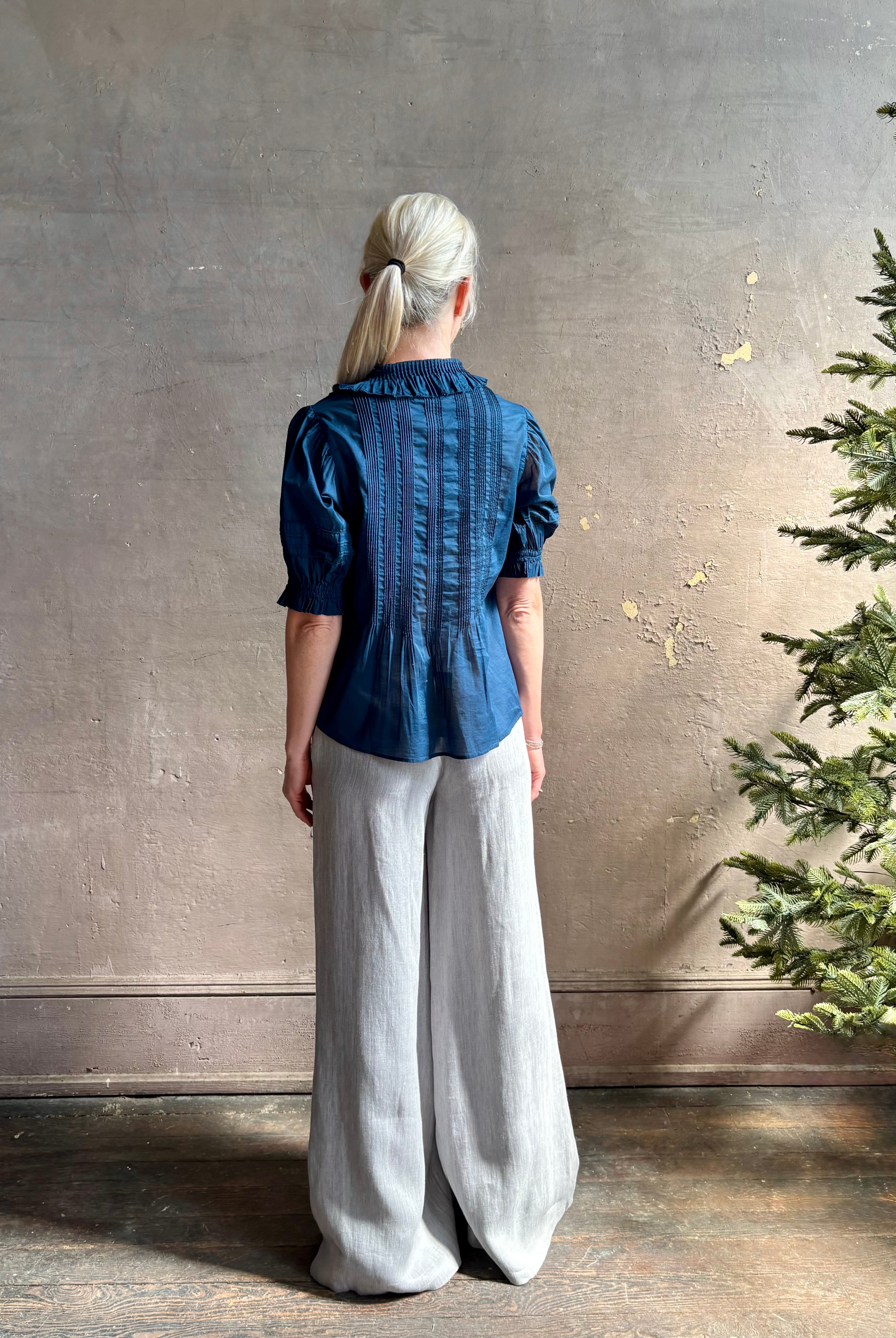 Mature woman wearing Jess Blouse from Trovata rendered in a classic Navy blue and crafted from light weight cotton poplin with short sleeves, peter pan collar, v-neck button front with intricate pintucking details at front and back