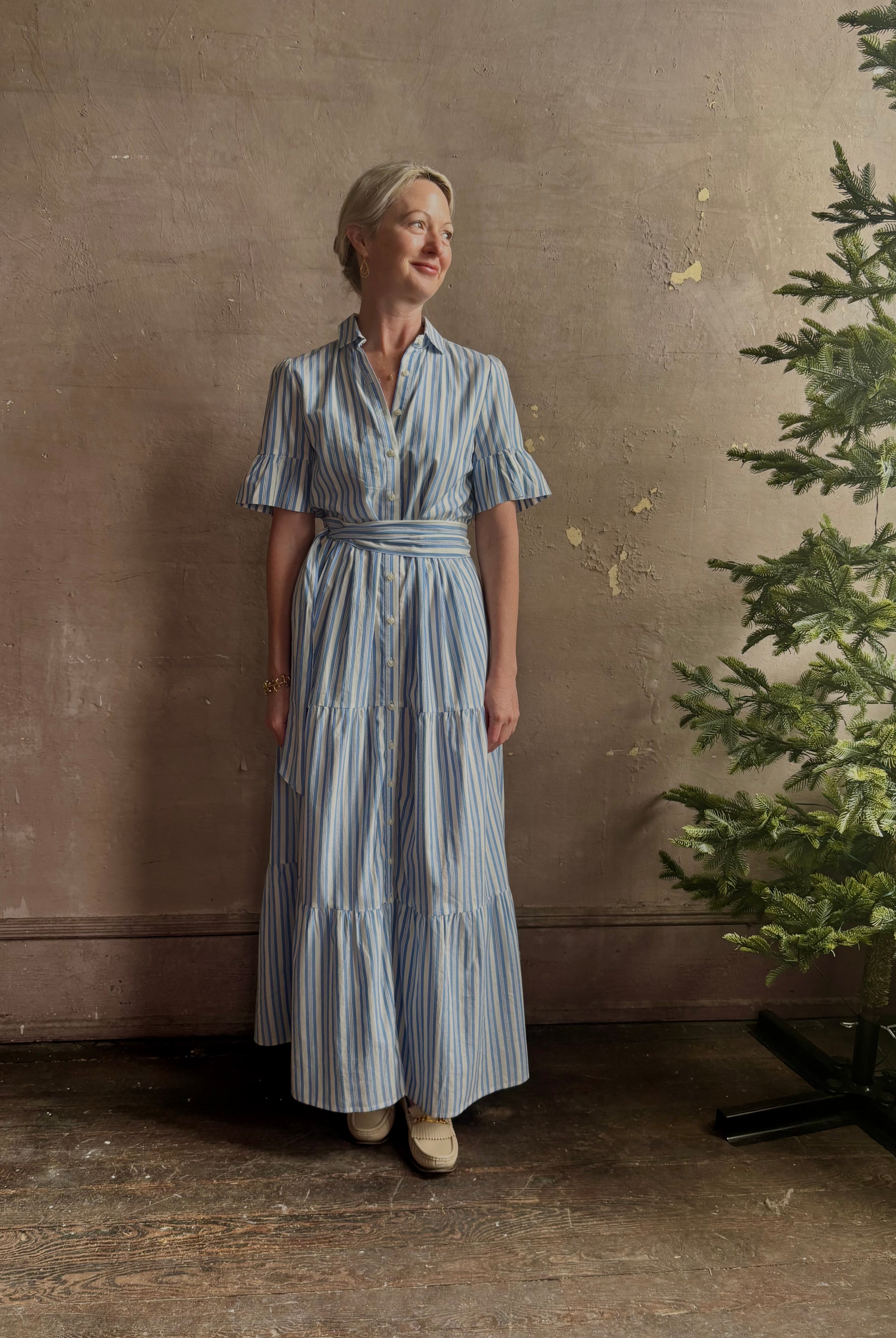 Mature woman wearing Martine maxi shirt Dress from Trovata rendered in a blue and white Bahia Stripe and crafted from cotton poplin with ruffle lined short sleeves, collar, side seam pockets, button front, tiered skirt and optional self tie co-ordinating fabric belt