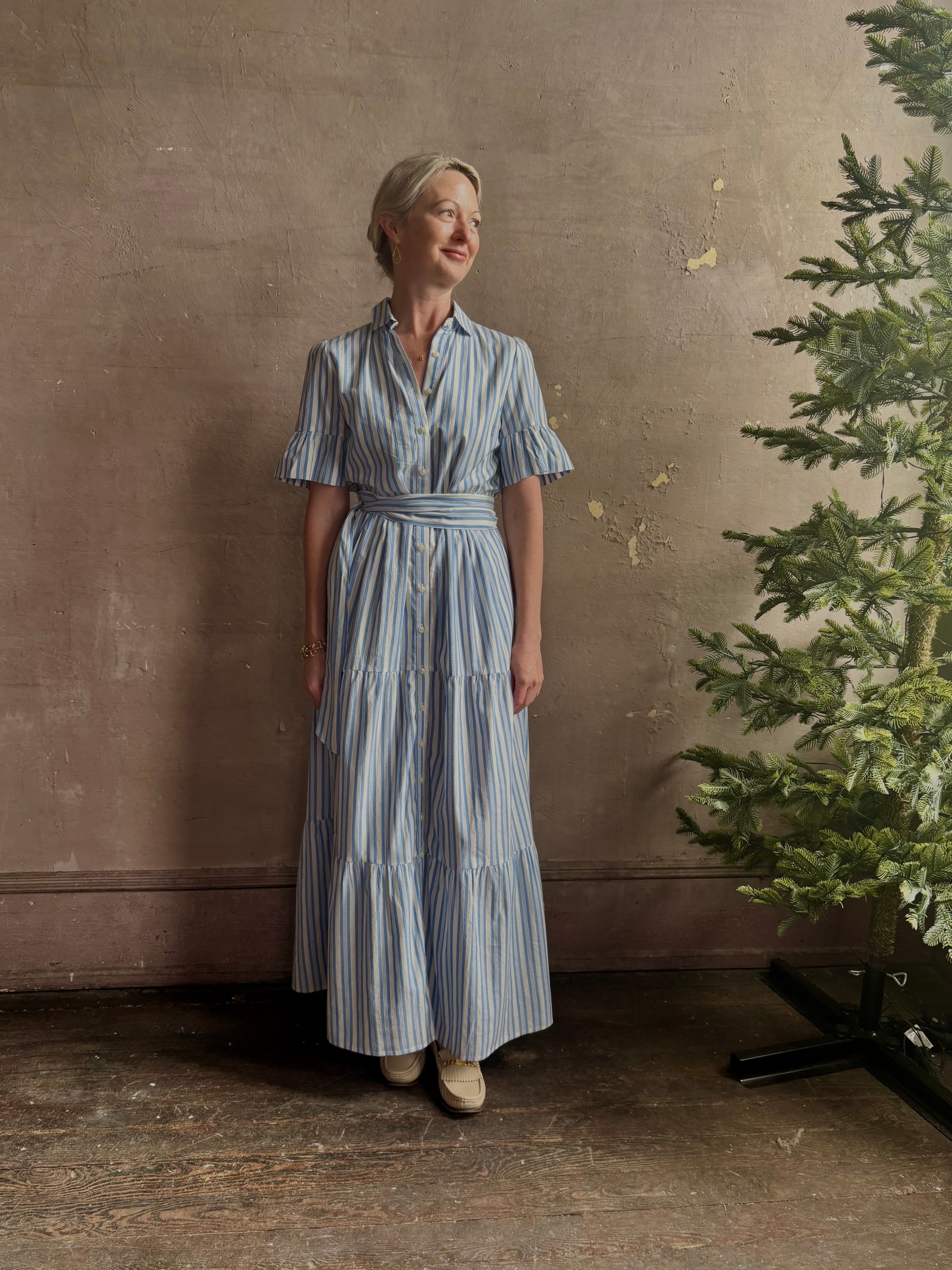 Mature woman wearing Martine maxi shirt Dress from Trovata rendered in a blue and white Bahia Stripe and crafted from cotton poplin with ruffle lined short sleeves, collar, side seam pockets, button front, tiered skirt and optional self tie co-ordinating fabric belt