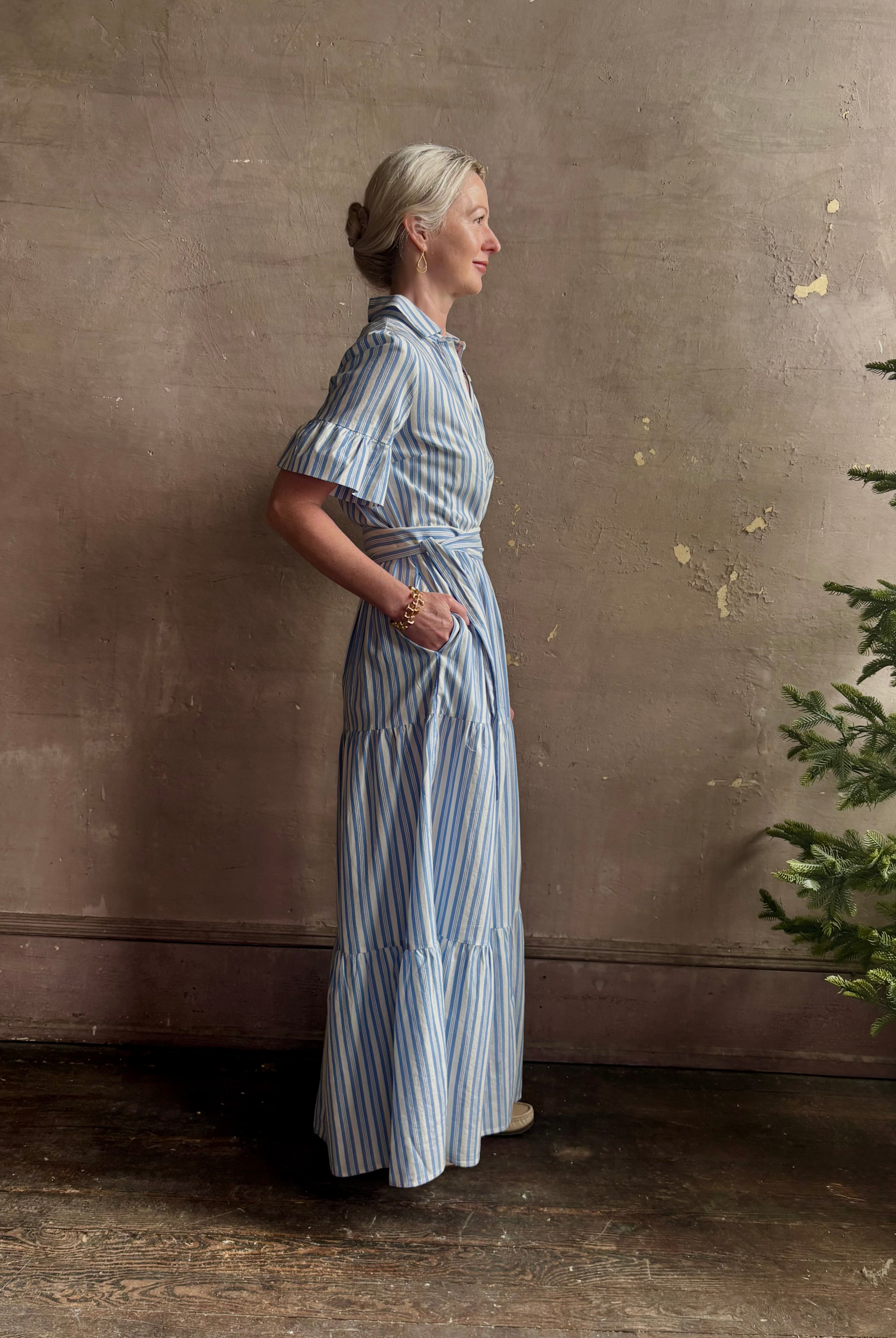 Mature woman wearing Martine maxi shirt Dress from Trovata rendered in a blue and white Bahia Stripe and crafted from cotton poplin with ruffle lined short sleeves, collar, side seam pockets, button front, tiered skirt and optional self tie co-ordinating fabric belt