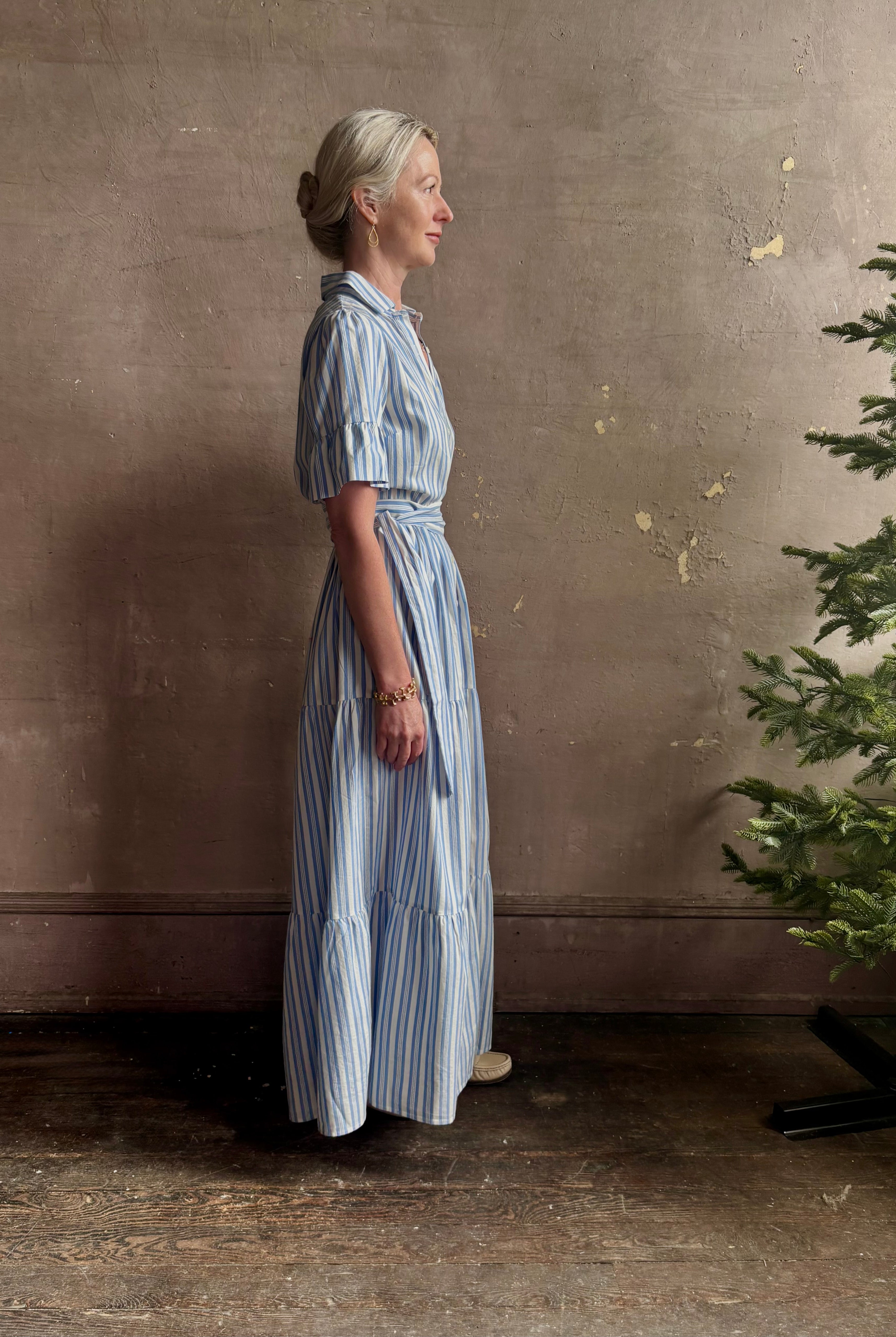 Mature woman wearing Martine maxi shirt Dress from Trovata rendered in a blue and white Bahia Stripe and crafted from cotton poplin with ruffle lined short sleeves, collar, side seam pockets, button front, tiered skirt and optional self tie co-ordinating fabric belt