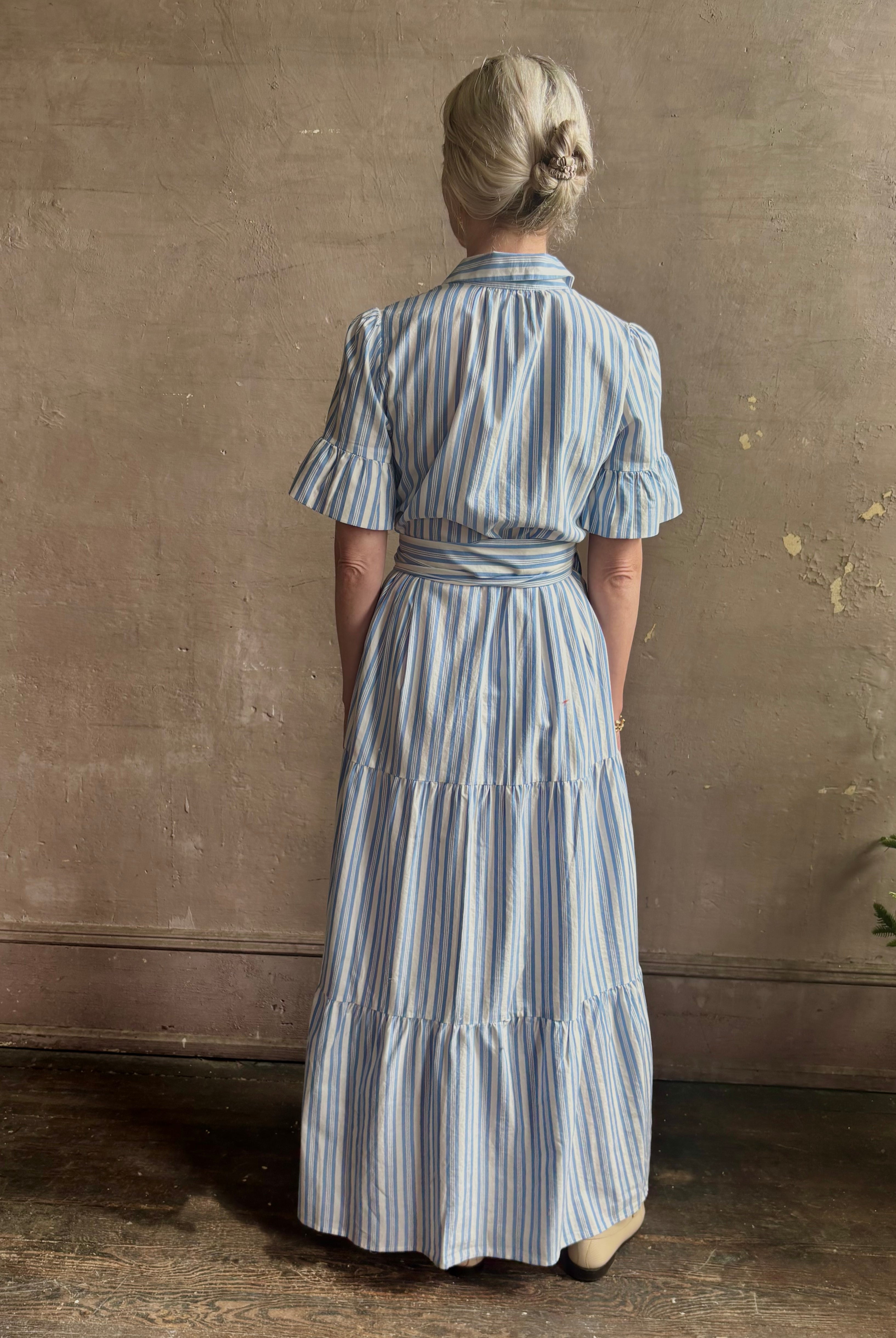 Mature woman wearing Martine maxi shirt Dress from Trovata rendered in a blue and white Bahia Stripe and crafted from cotton poplin with ruffle lined short sleeves, collar, side seam pockets, button front, tiered skirt and optional self tie co-ordinating fabric belt