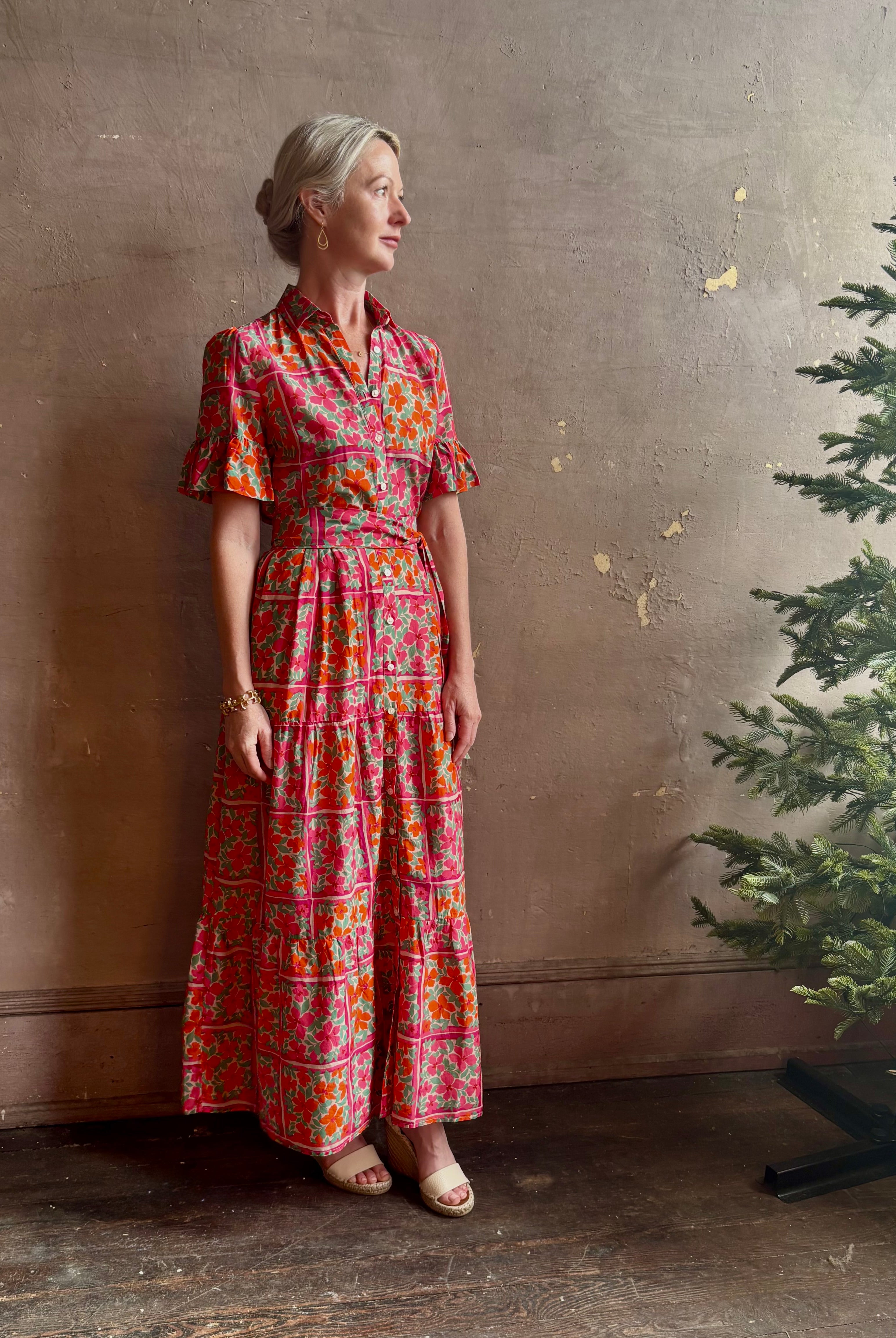 Mature woman wearing Martine maxi Dress from Trovata rendered in the Botanic Tile pink and orange floral print and crafted from a luxurious blend of cotton and silk with button front, collar, short sleeves and matching optional self tile belt